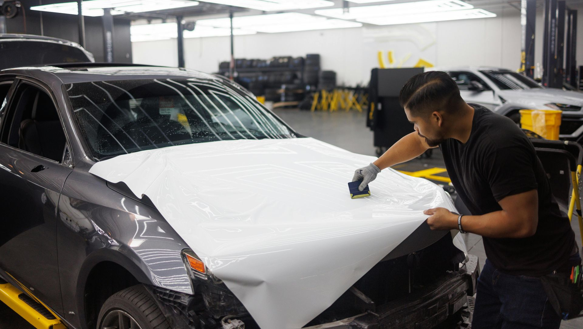 Two men applying white vinyl wrap to a gray car hood inside a workshop.