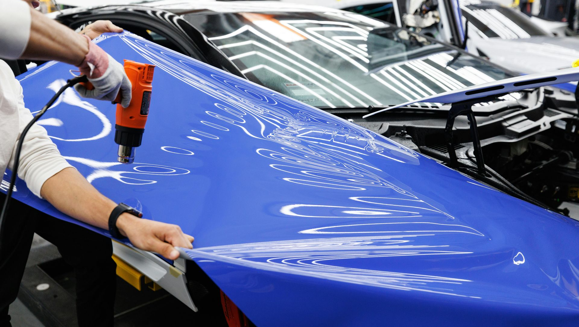 Person applying blue vinyl wrap to a car's hood with a heat gun in a shop.