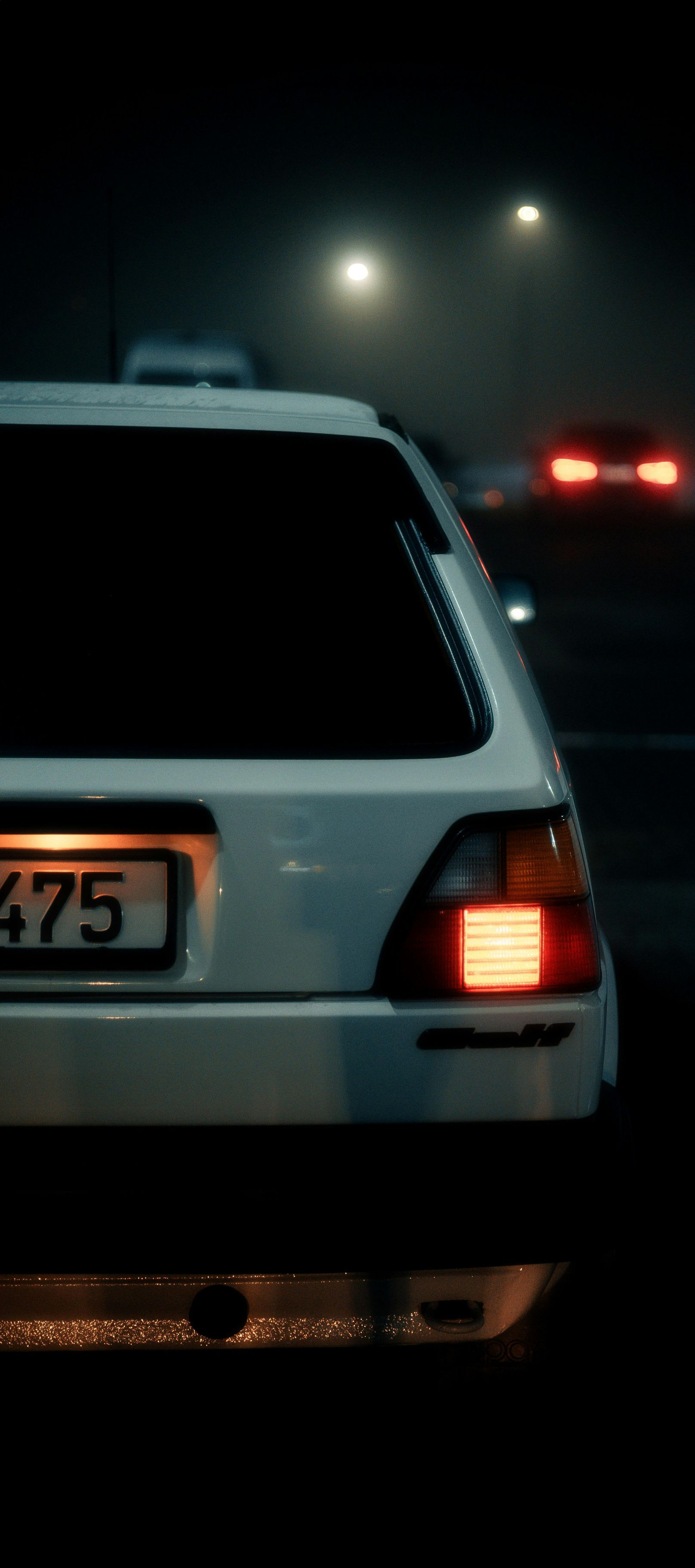 White car's rear view in the dark, with taillight illuminated. Other cars and streetlights blurred in background.