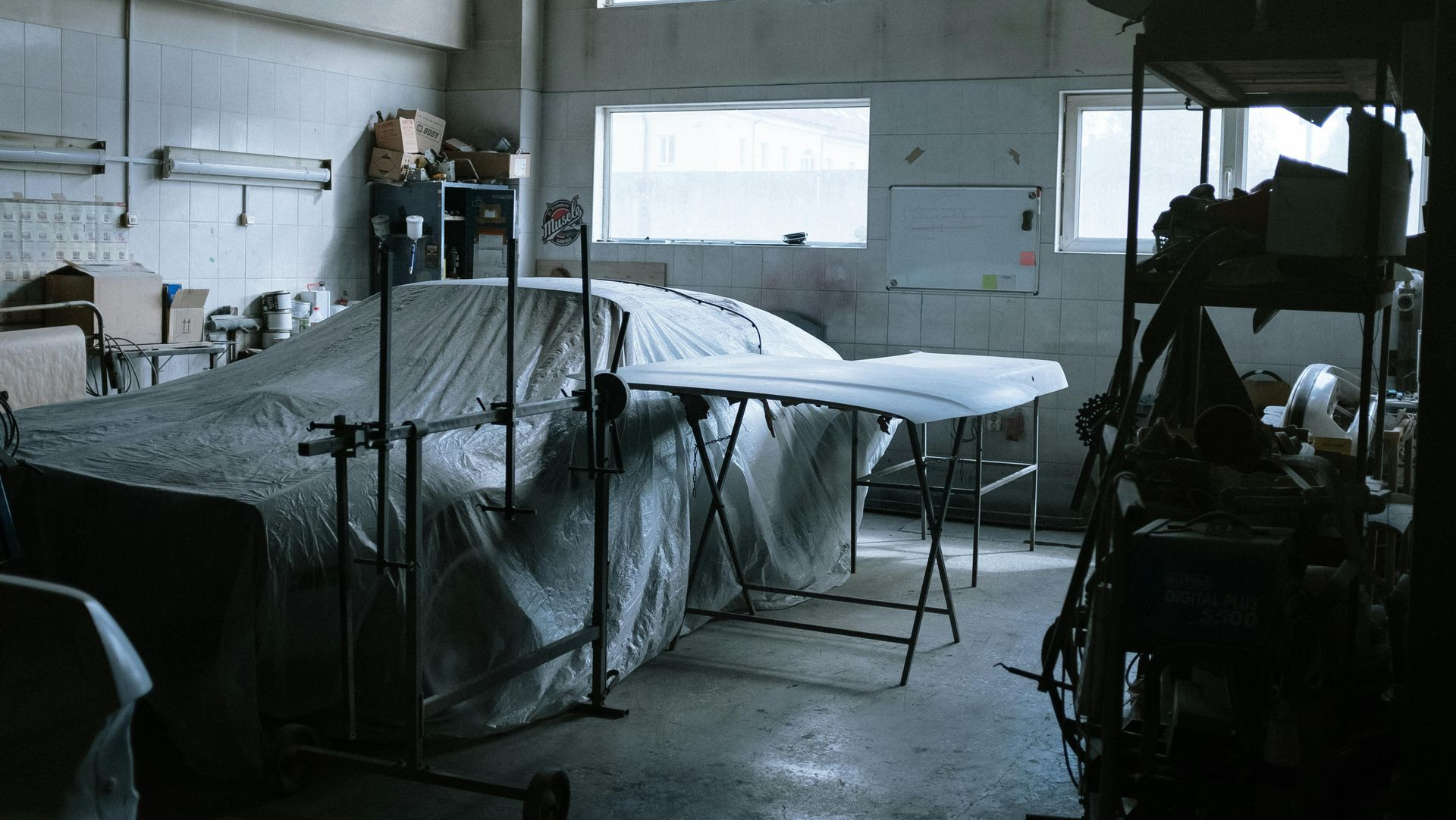 Car repair shop interior with covered car and hood; workbench, shelves, and windows visible.