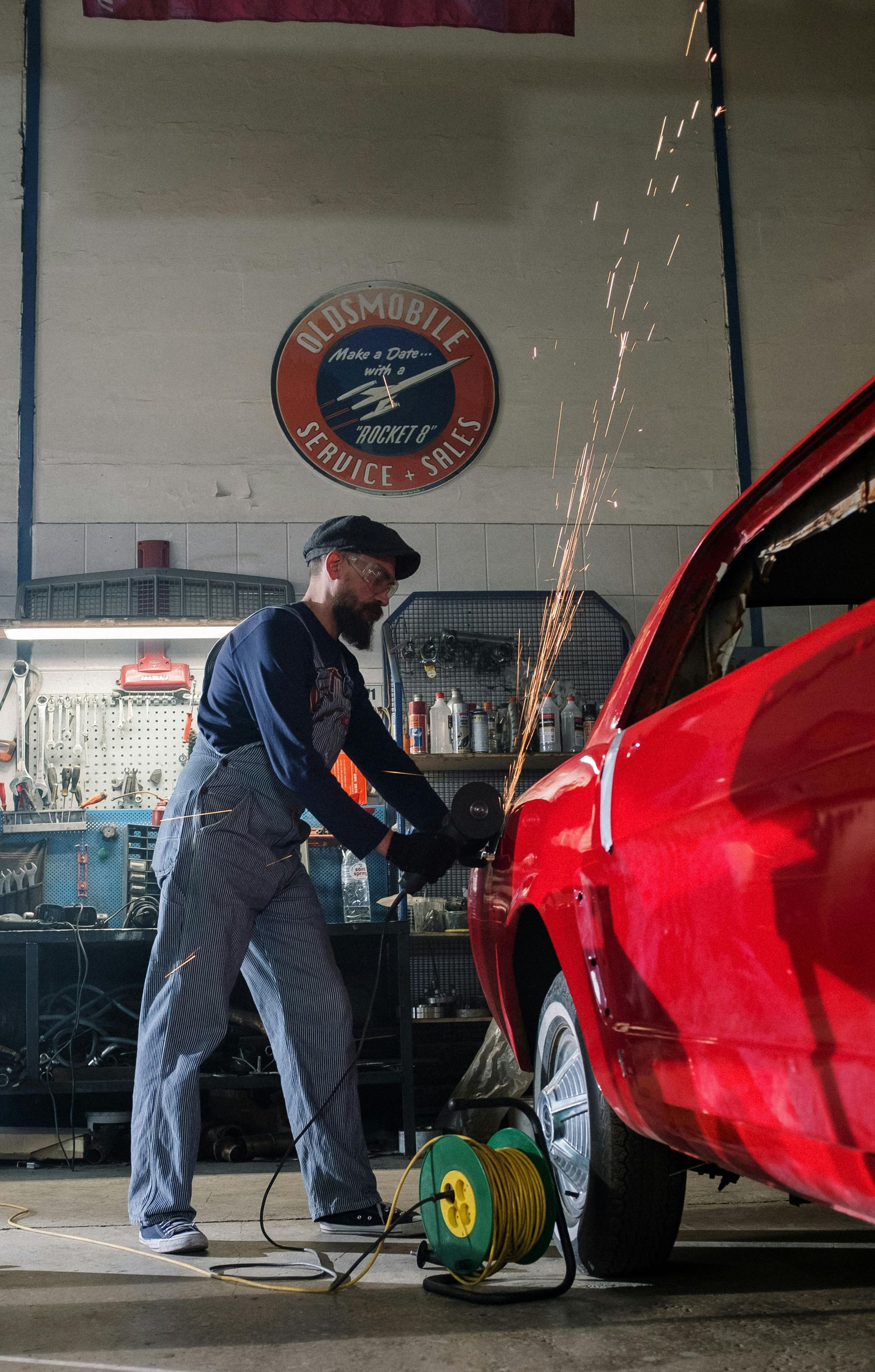 Mechanic in striped overalls using a grinder on a red car in a garage, sparks flying.