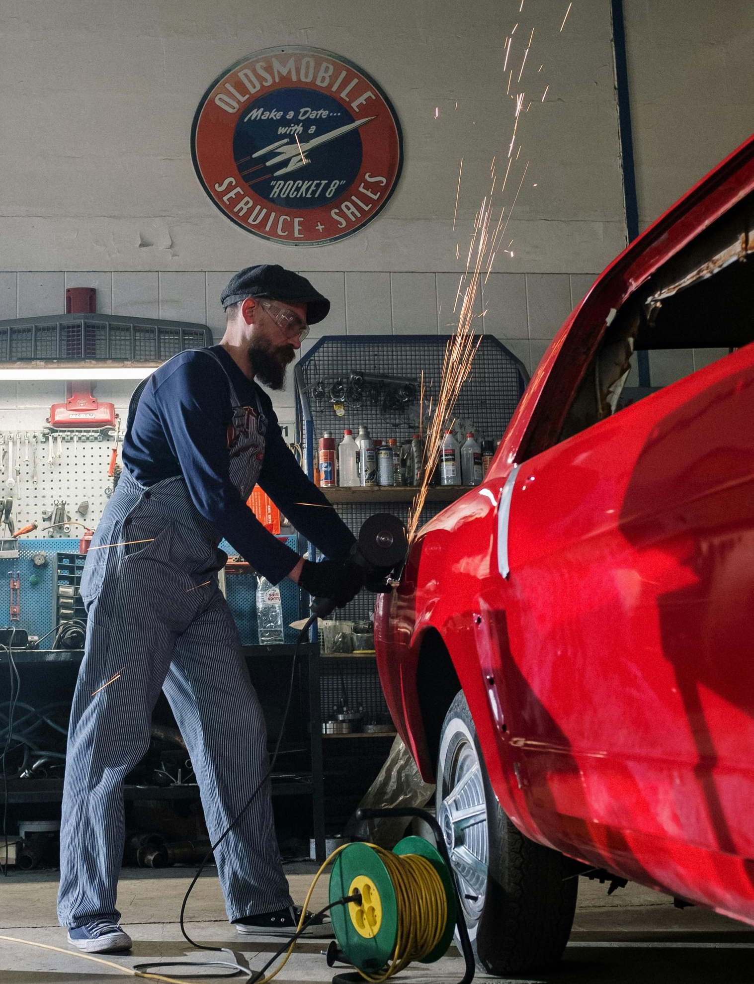Man in overalls using a grinder on a red car in a garage, sparks flying.