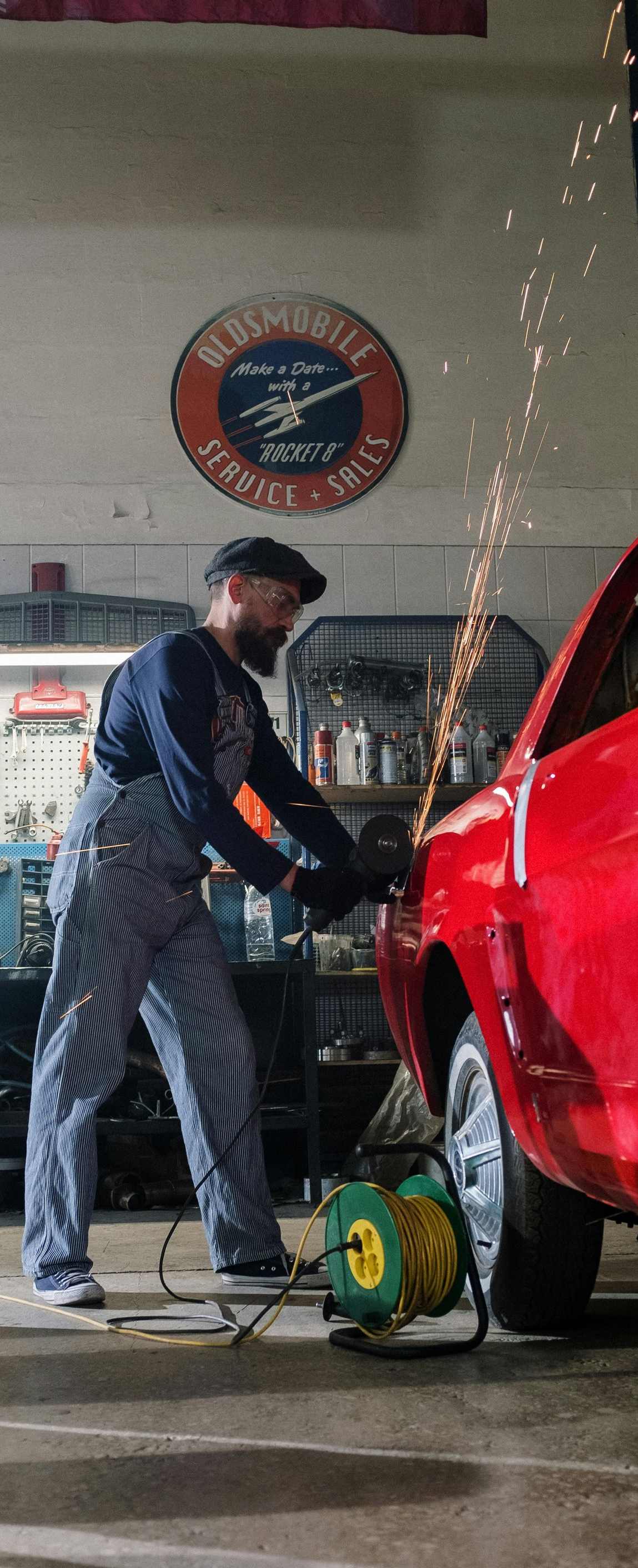 Mechanic in striped overalls using a grinder on a red car in a garage, sparks flying.