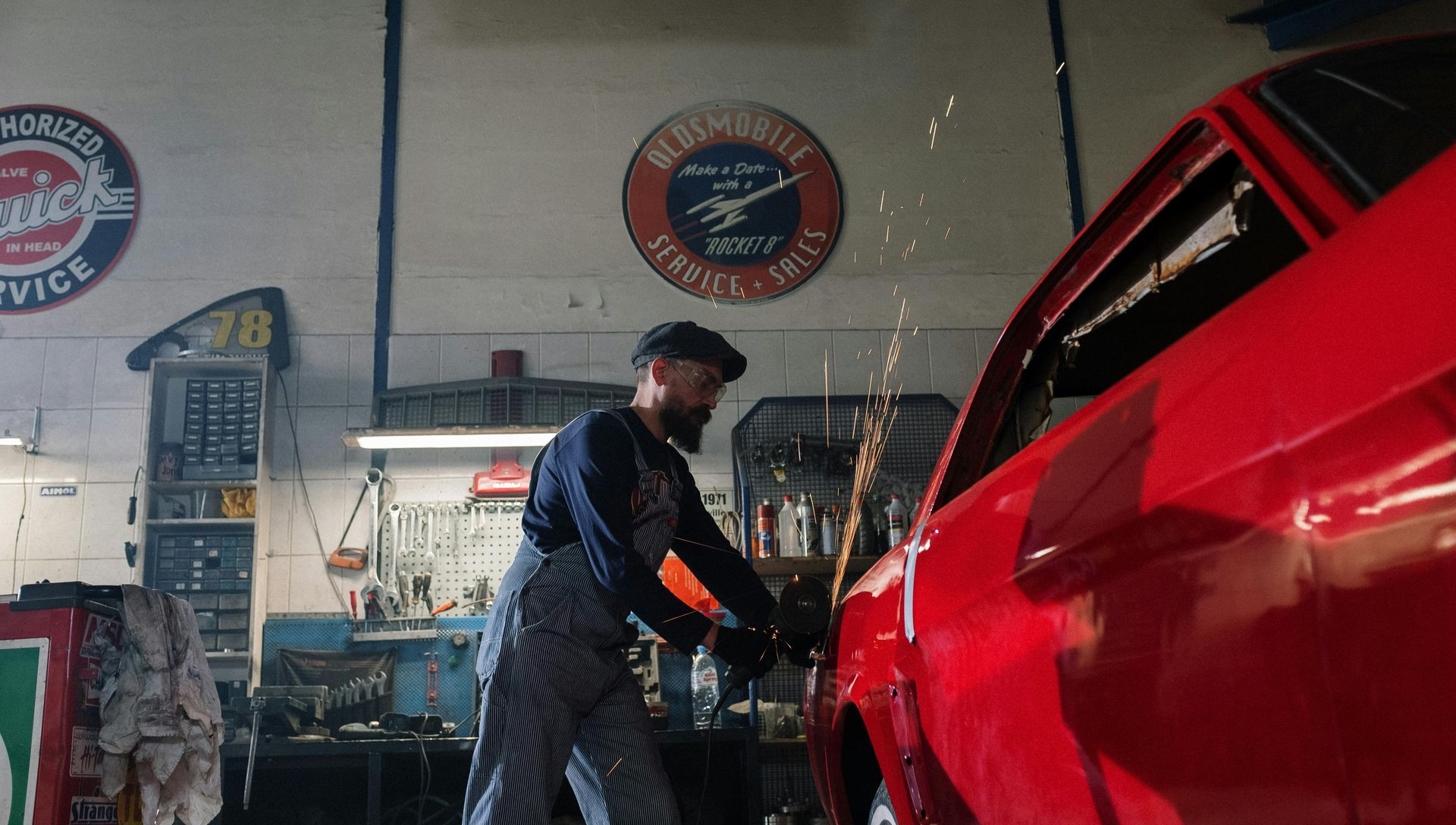 Man in overalls using a grinder on a red car in a garage, sparks flying.