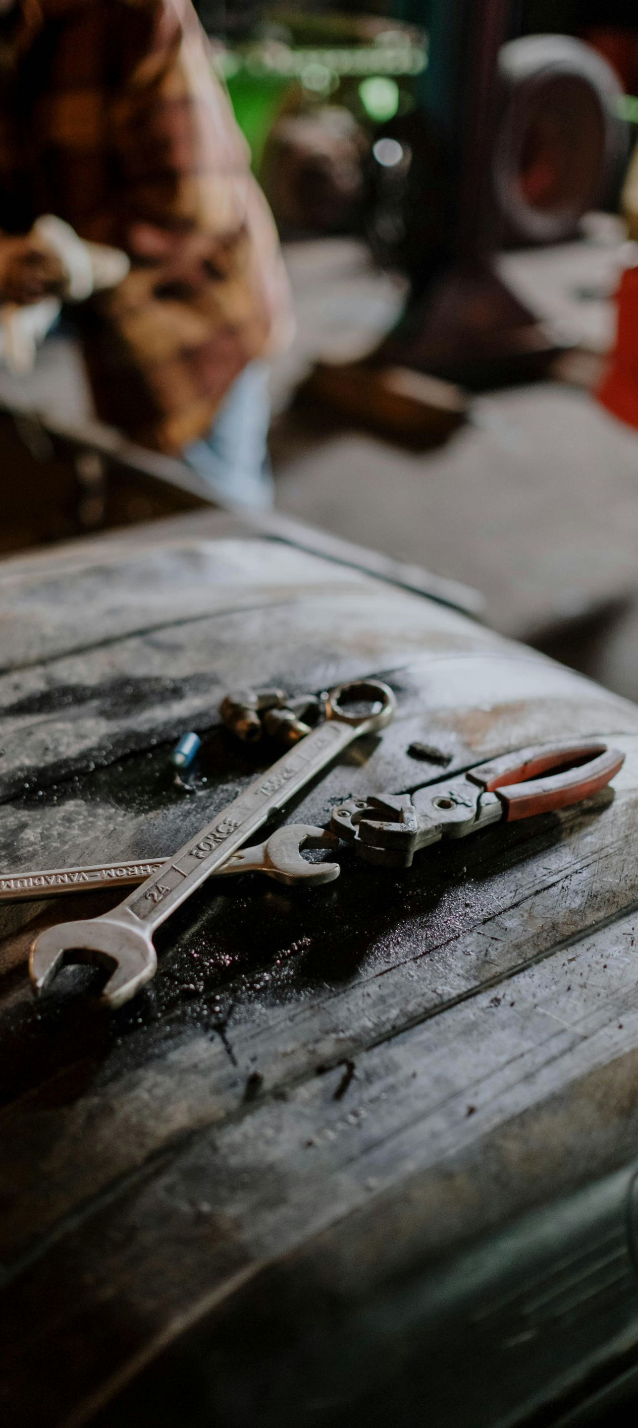 Wrench and pliers resting on a dark, textured surface, possibly a vehicle part; person out of focus in the background.