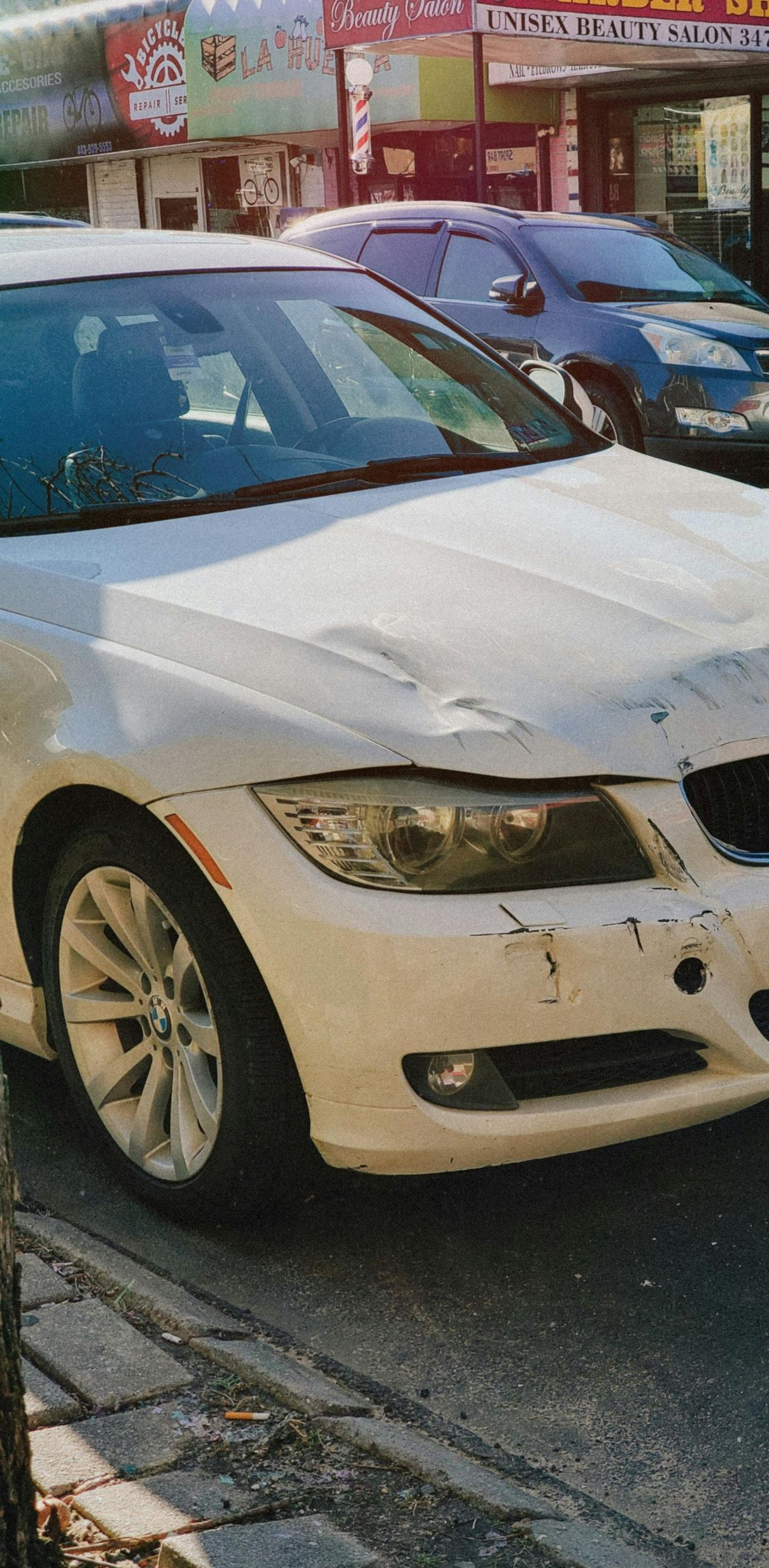 White BMW with front-end damage parked on a city street. Other cars and storefronts are visible in the background.