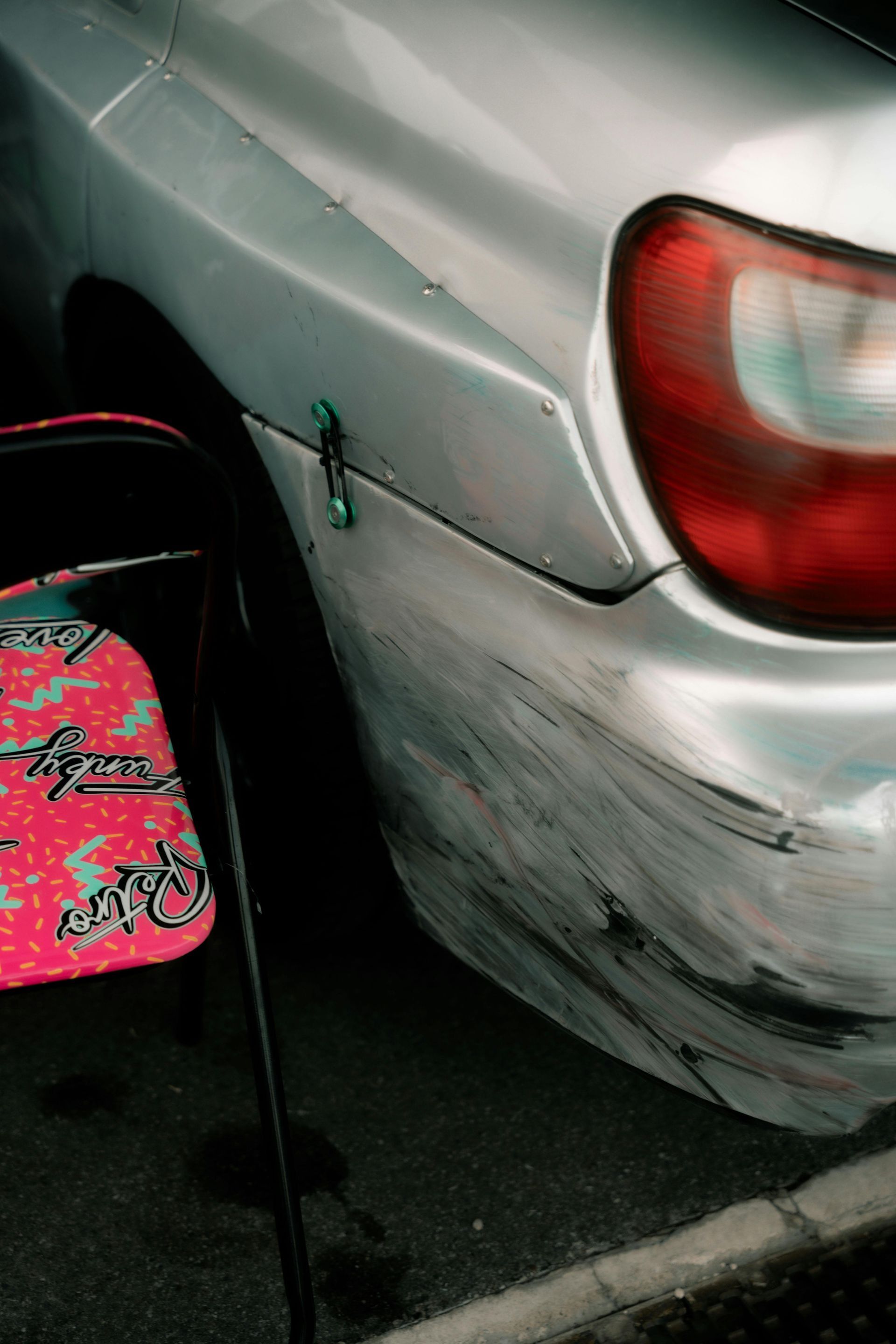 A damaged silver car bumper with heavy scuff marks, positioned next to a pink patterned folding chair.