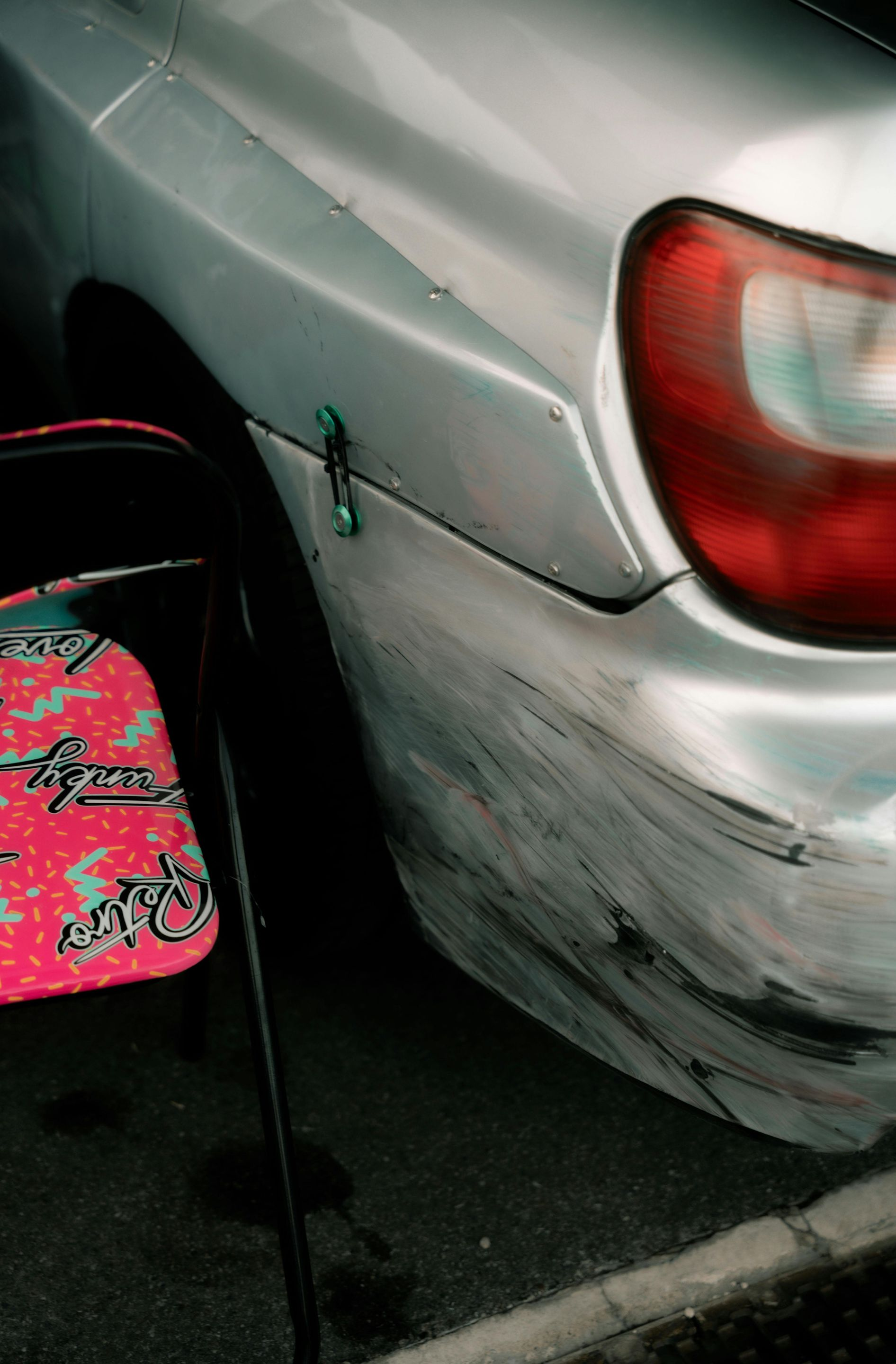 A damaged silver car bumper with heavy scuff marks, positioned next to a pink patterned folding chair.