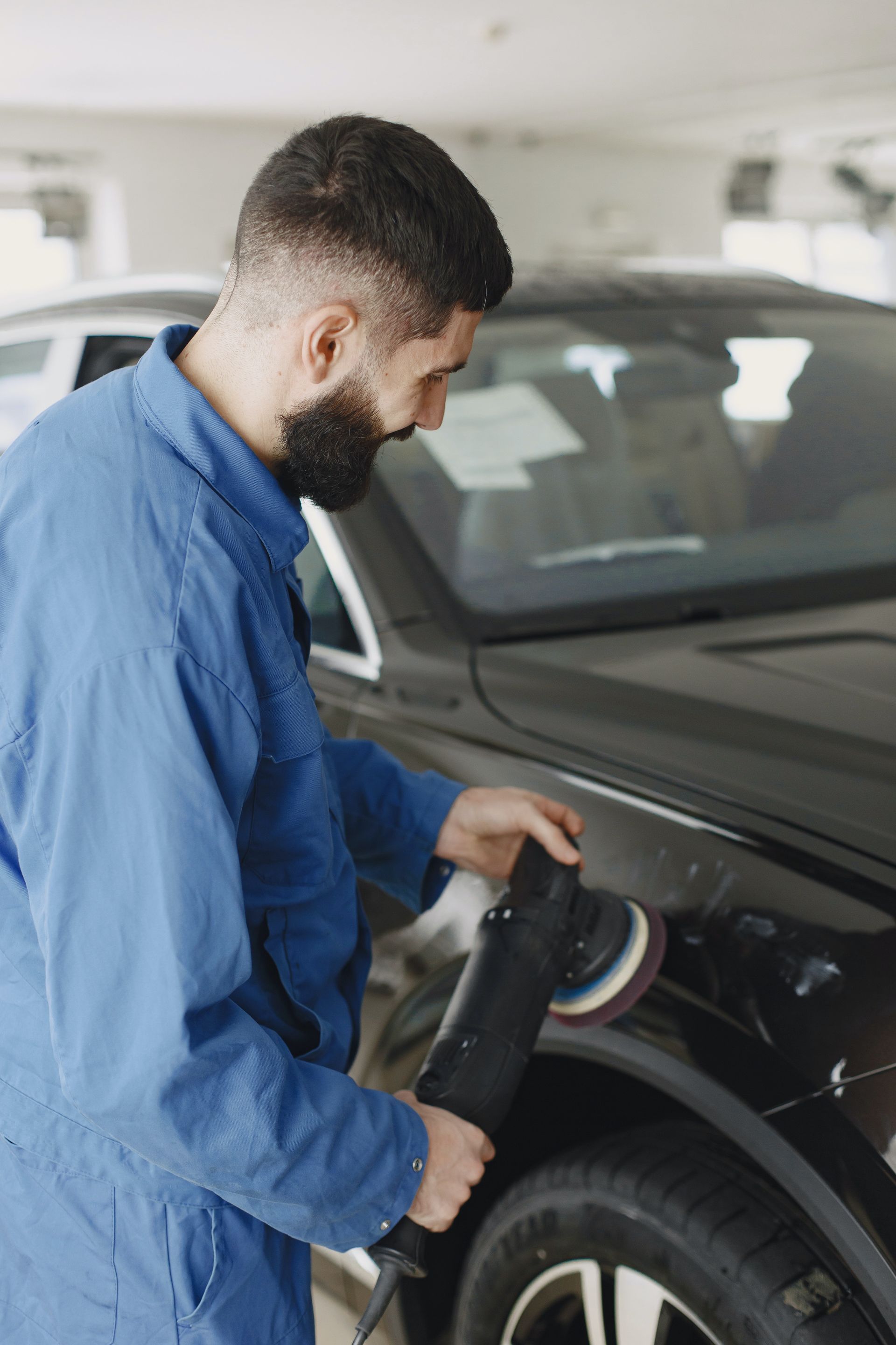 A man in a blue jumpsuit polishes a black car with a power buffer in a garage.