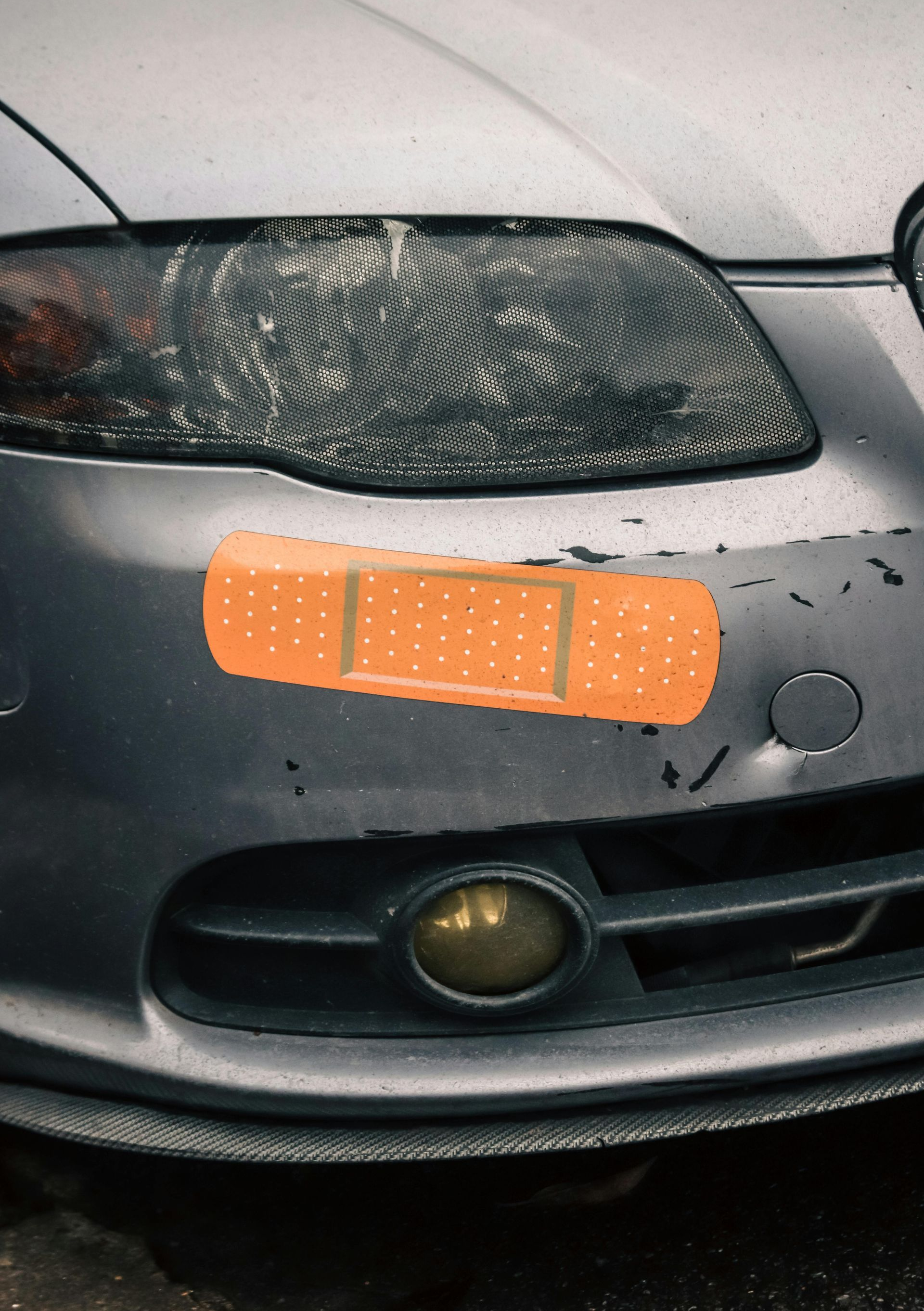 A large, orange adhesive bandage is placed over chipped paint on the gray front bumper of a car.