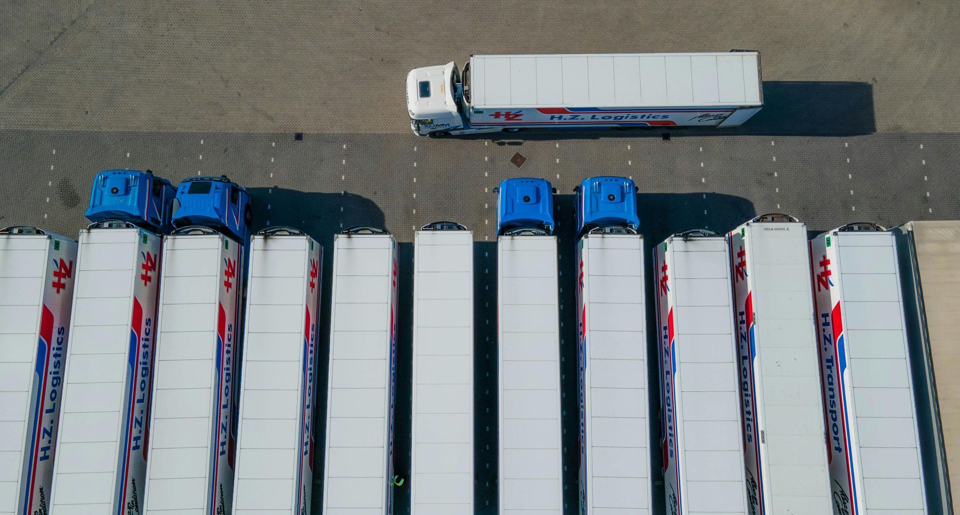 Overhead view of several white semi-truck trailers with blue cabs parked in a lot.