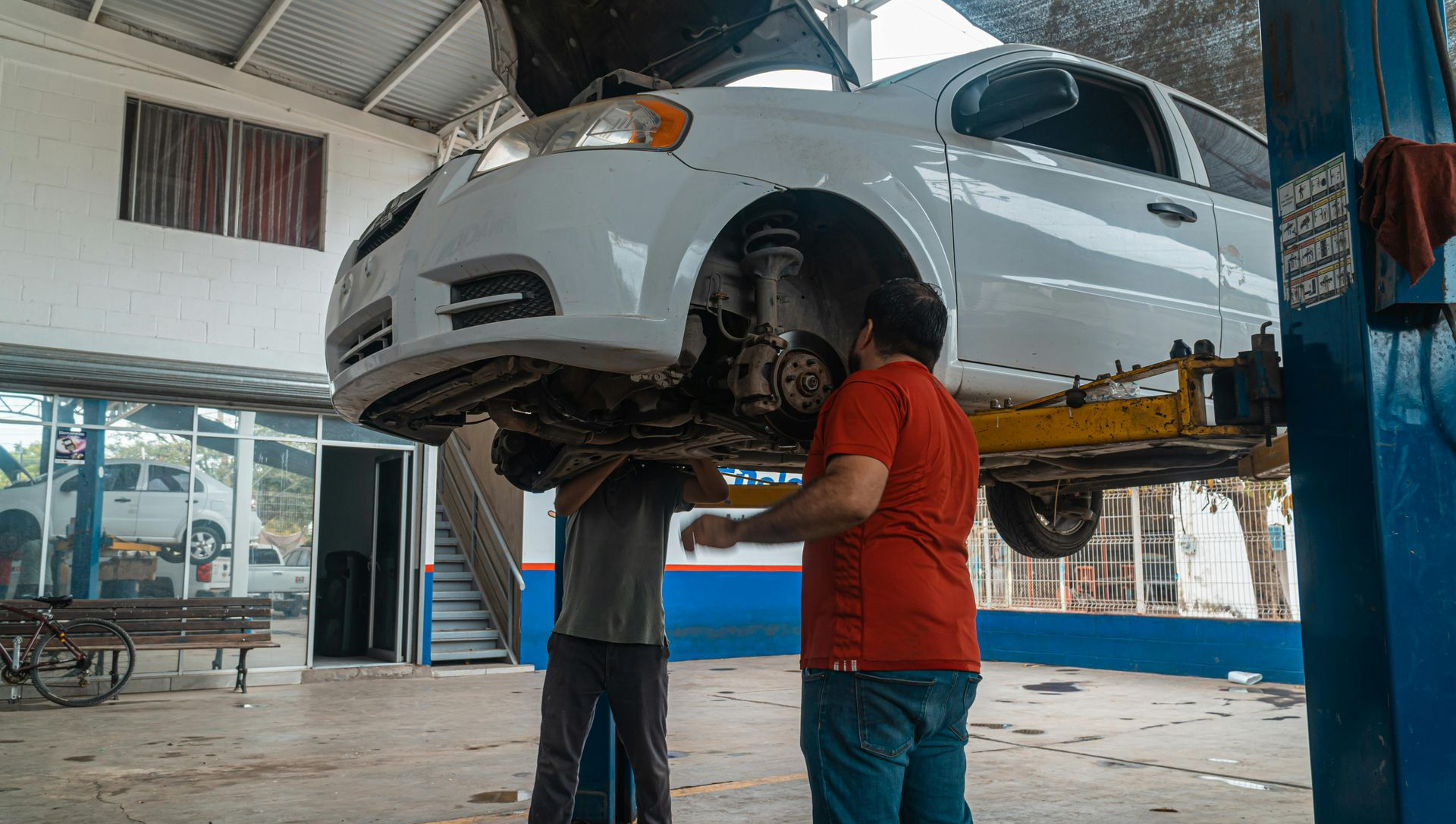 Two mechanics working on a white car raised on a lift in a repair shop.