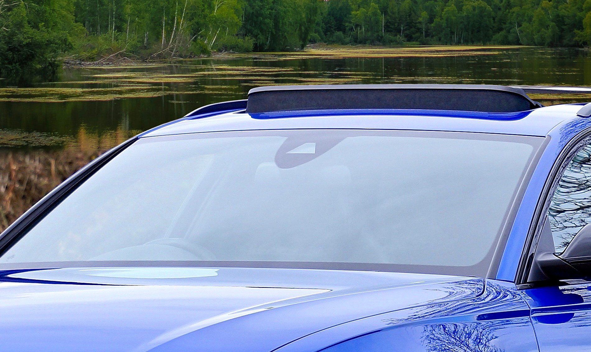 Blue car's windshield with a black sensor housing on the roof, set against a lake and trees.