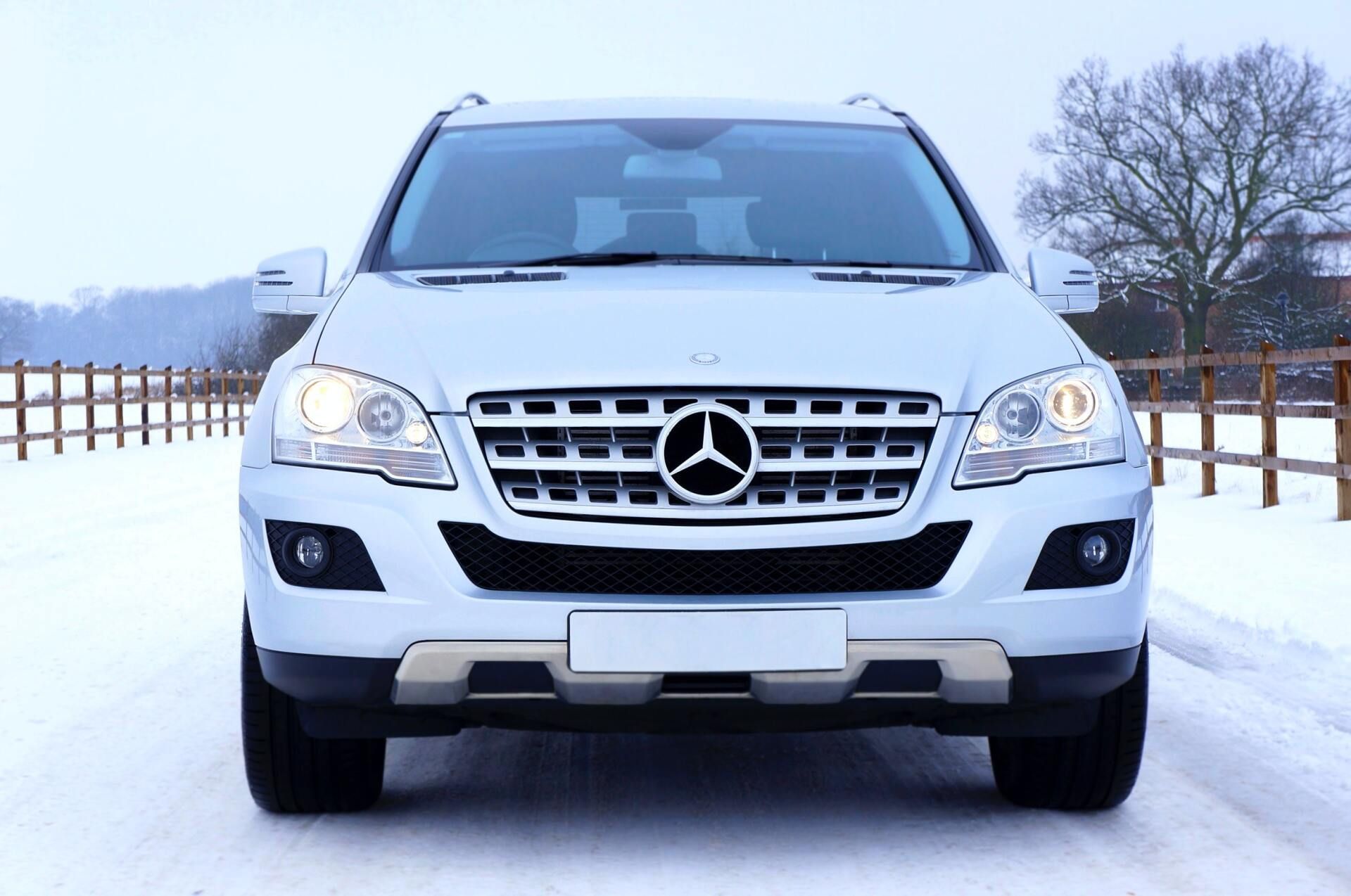 Silver Mercedes SUV on a snowy road, facing the camera. The sky is overcast and a wooden fence lines the sides.