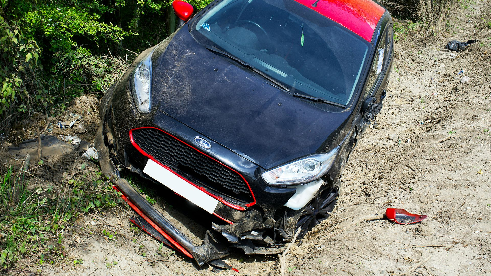 A black Ford hatchback with a red roof sits in a ditch with front-end damage near a dirt path and green foliage.