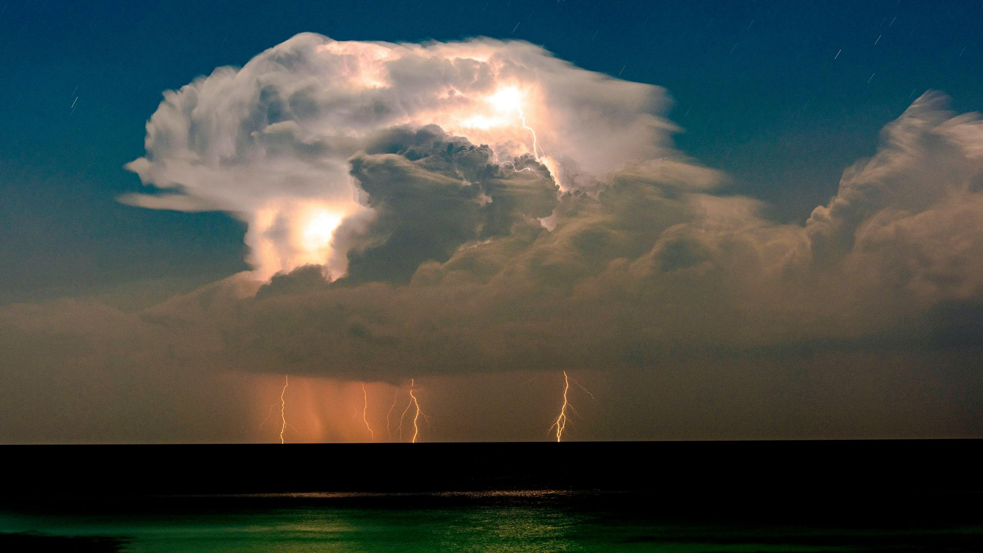 Lightning illuminates a large storm cloud over dark water; streaks of rain and lightning visible.