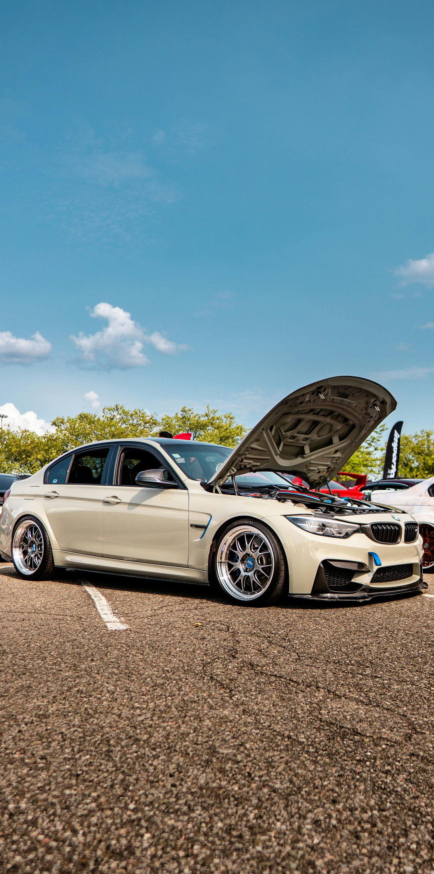 White BMW sedan with open hood, parked on asphalt, bright blue sky.