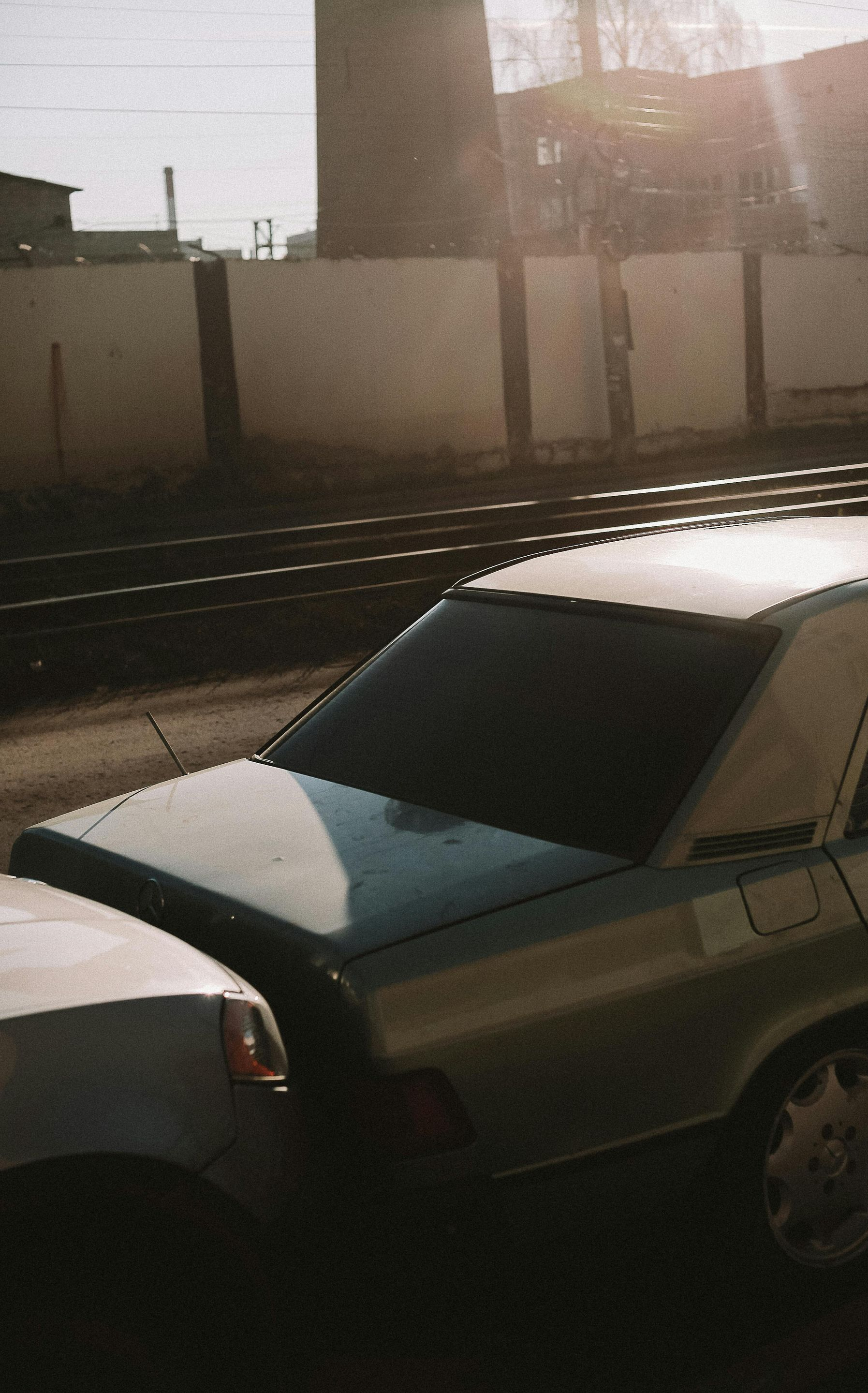 A silver sedan parked on a street near a fenced industrial area under bright sunlight.