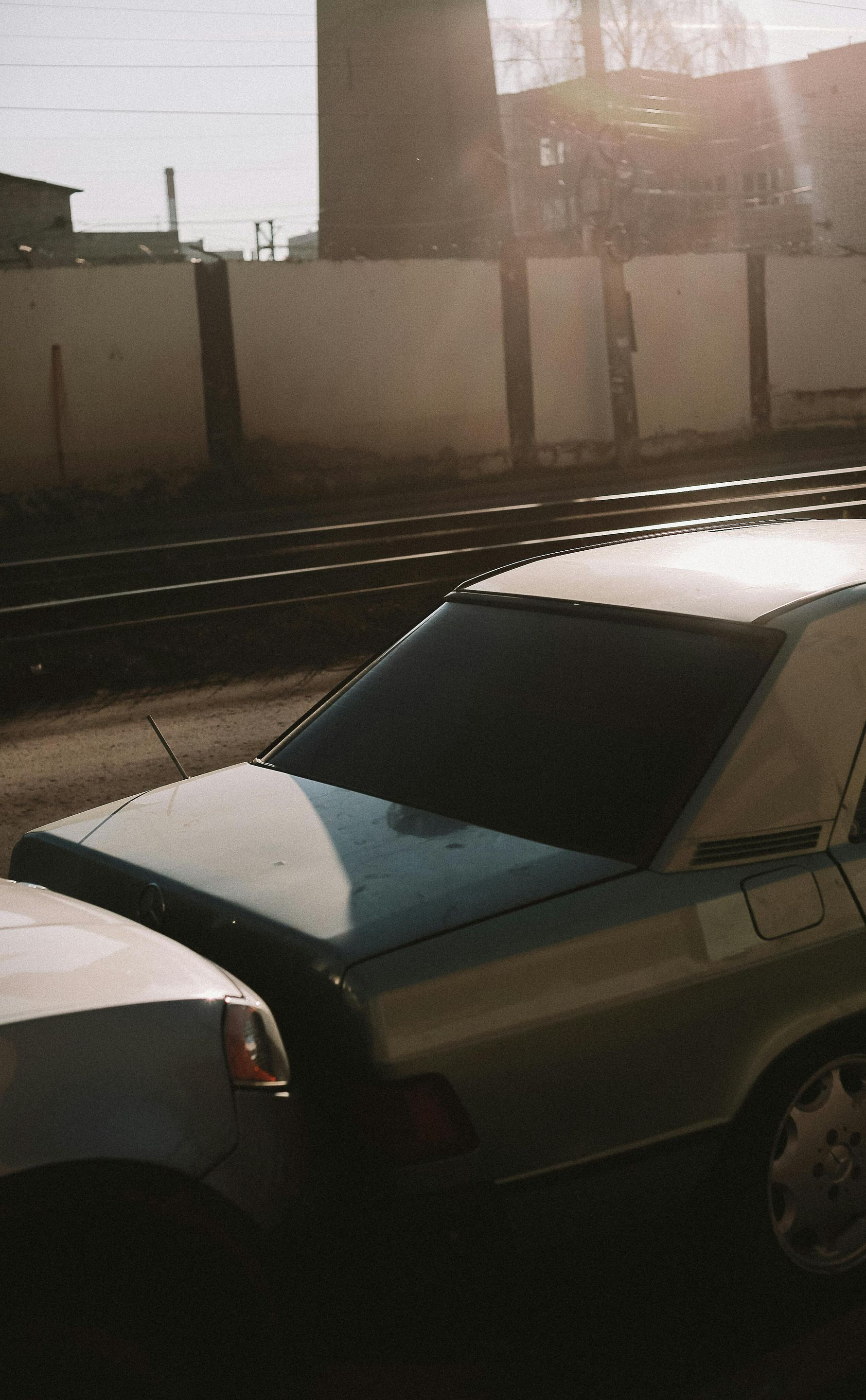 Two cars parked next to each other by railroad tracks with a brick building in the background.