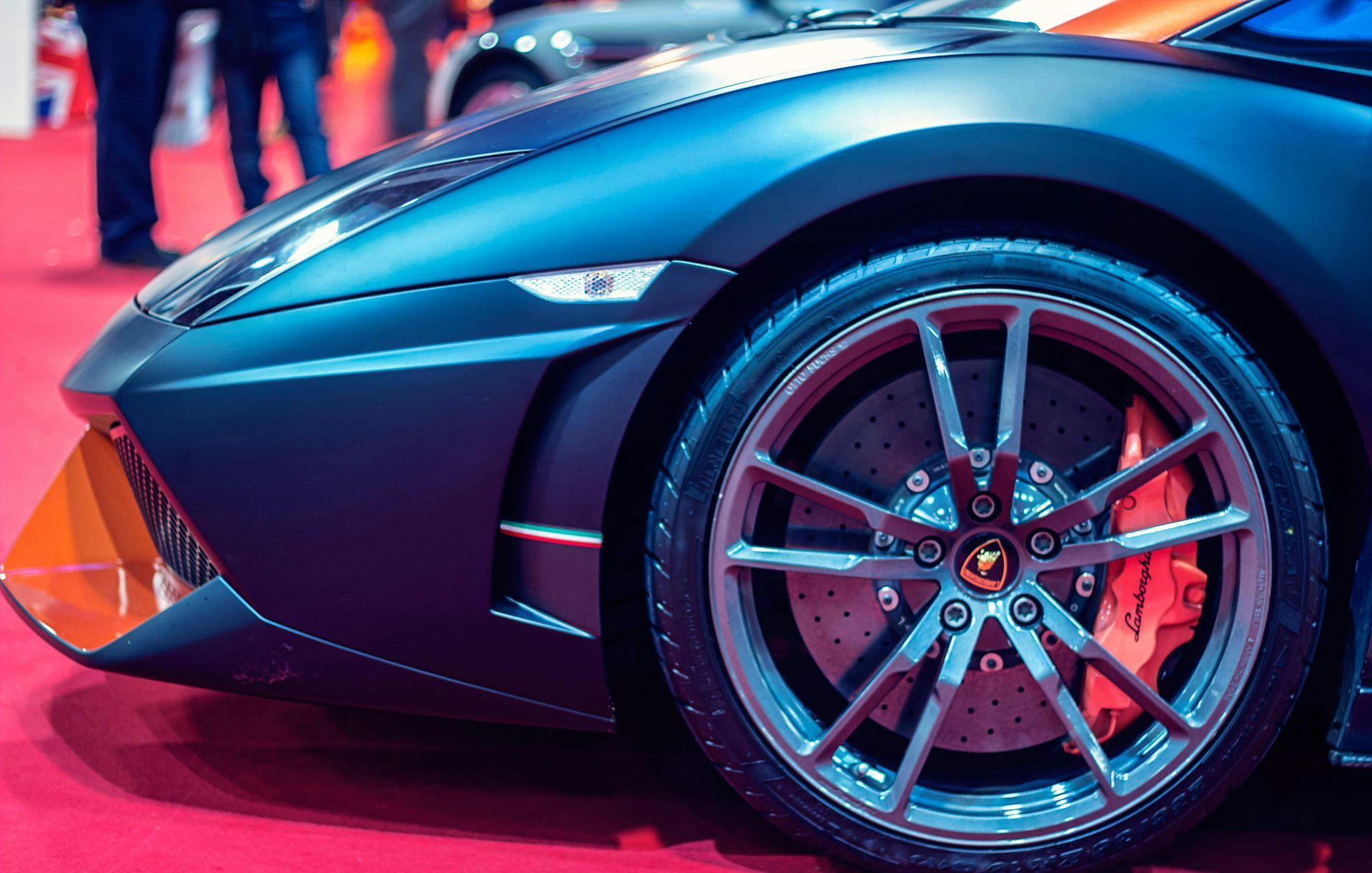 Close-up of a dark blue Lamborghini sports car's front wheel. The wheel has silver spokes, with red brake calipers and a red carpet background.
