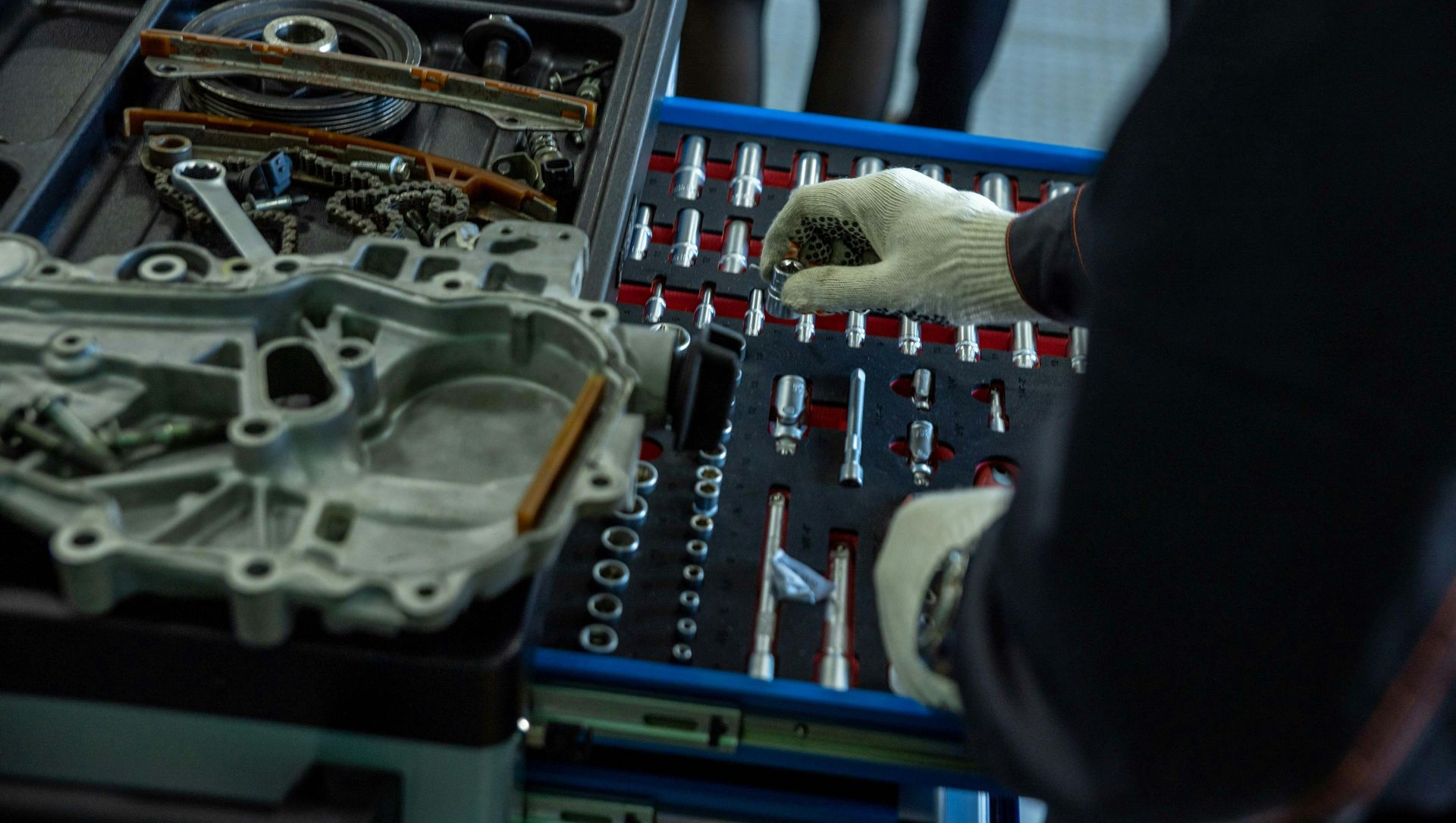 Mechanic's gloved hand selecting a tool from a tray of sockets and wrenches, near a disassembled engine and parts.