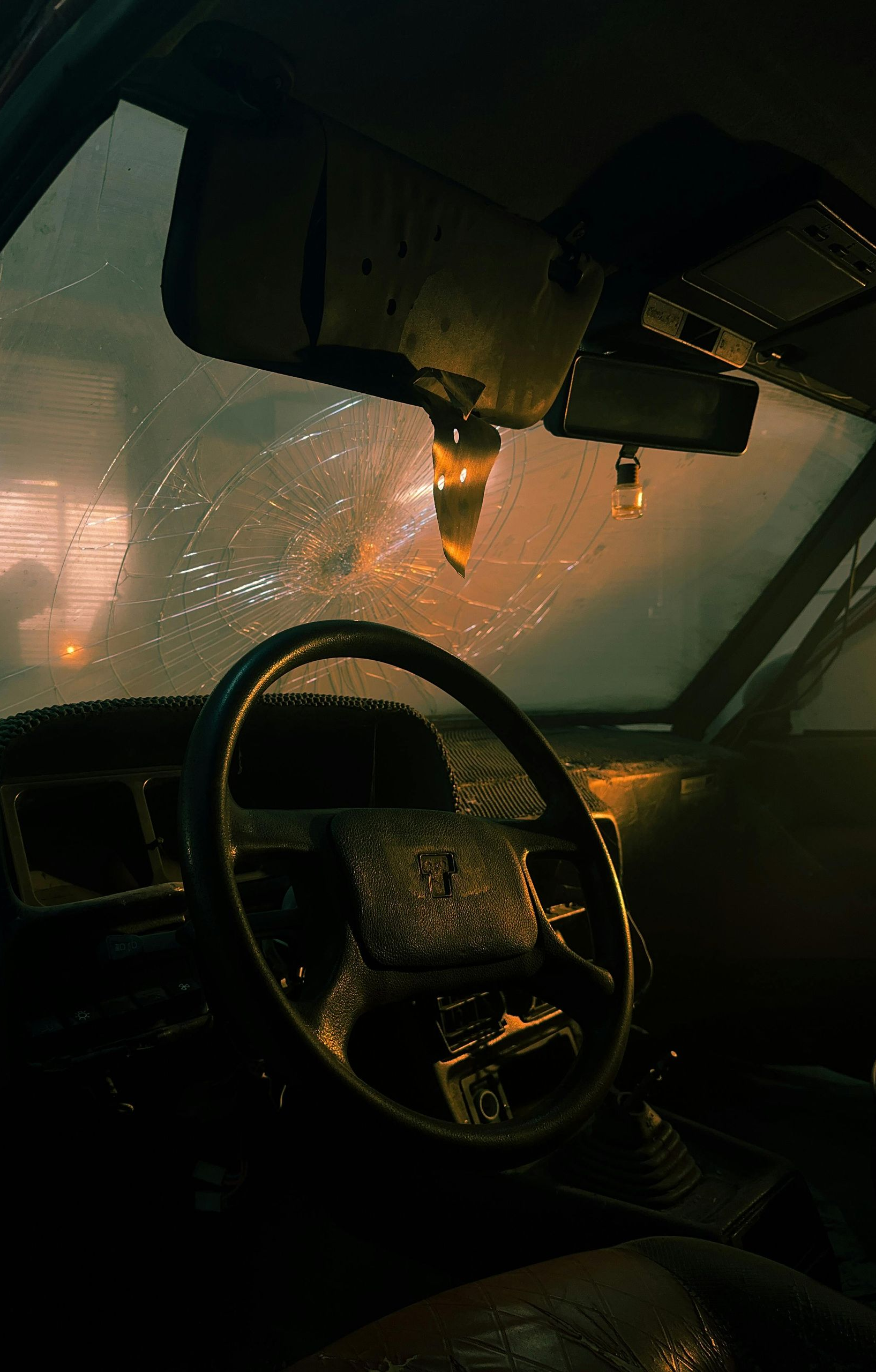 Interior of a wet car, steering wheel in focus, windshield speckled with rain, illuminated by warm orange light.