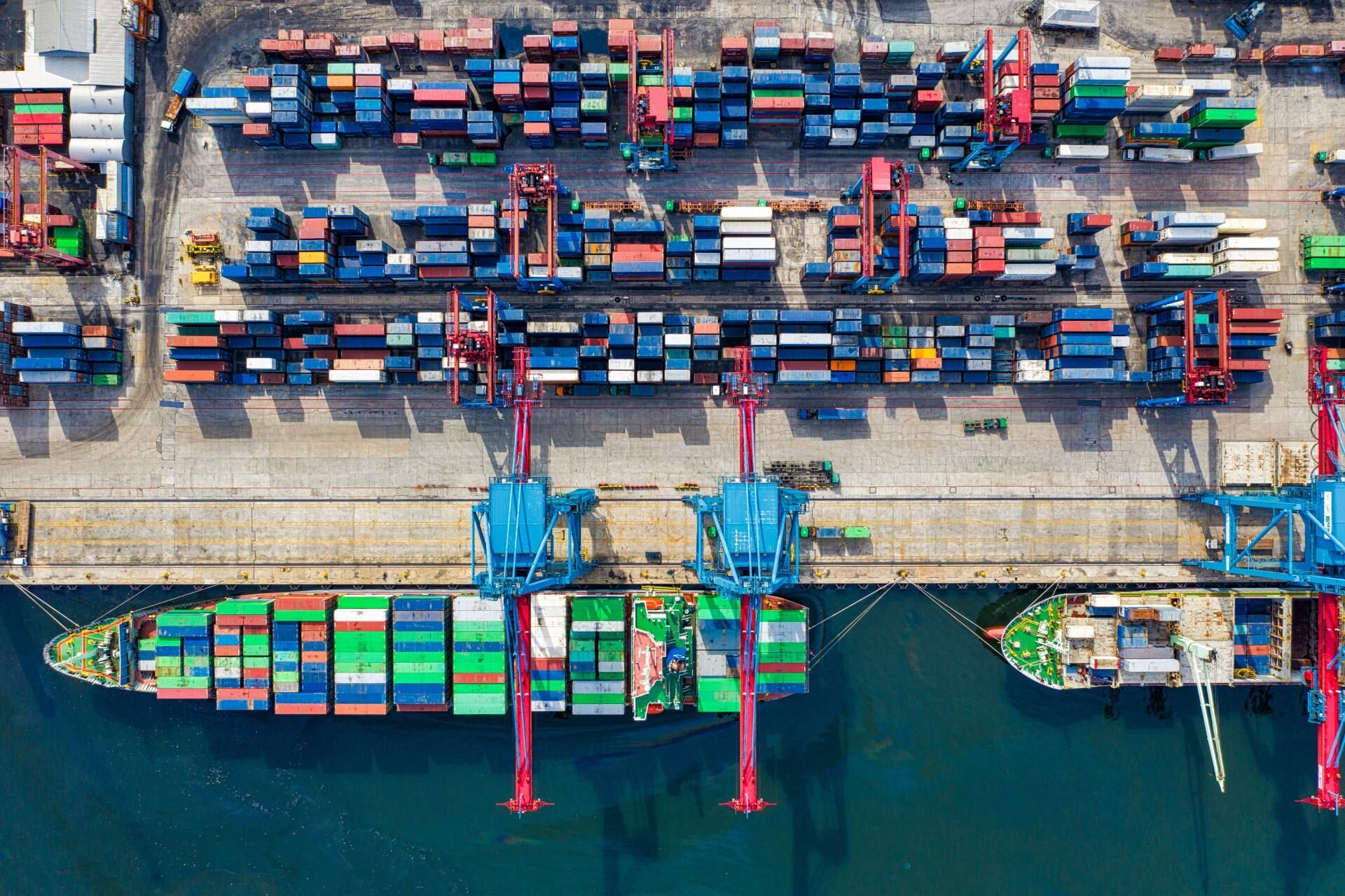 Aerial view of a busy shipping port with container ships, colorful stacked cargo containers, and cranes in operation.