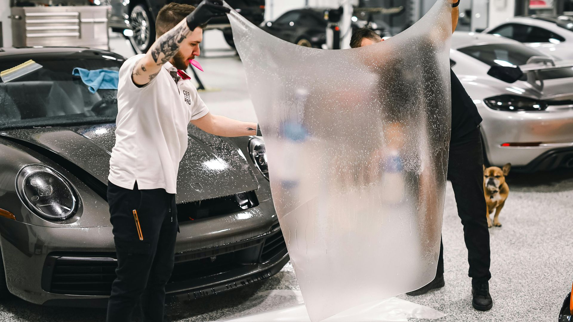 Two people applying protective film to a dark gray Porsche in a garage.