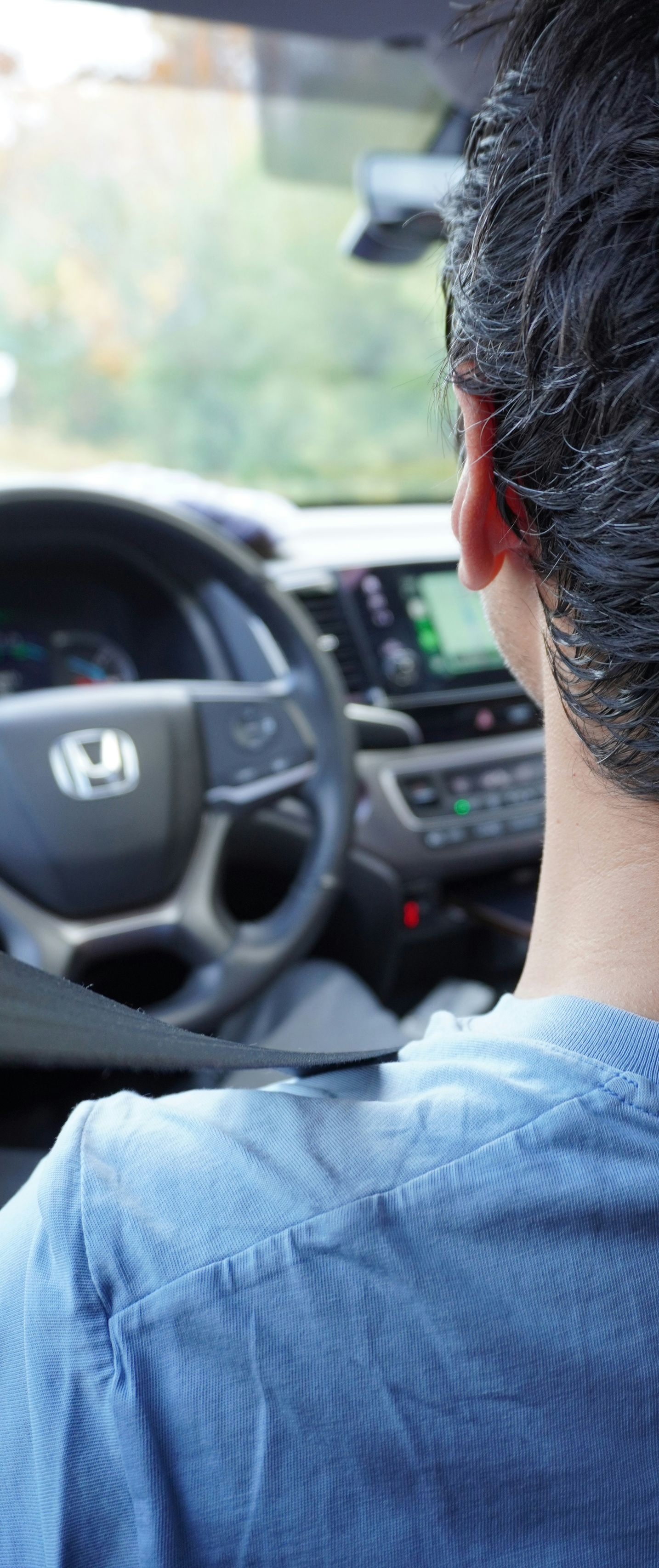 Person driving a car, viewed from behind. Steering wheel, dashboard, and outside view visible. Blue shirt.