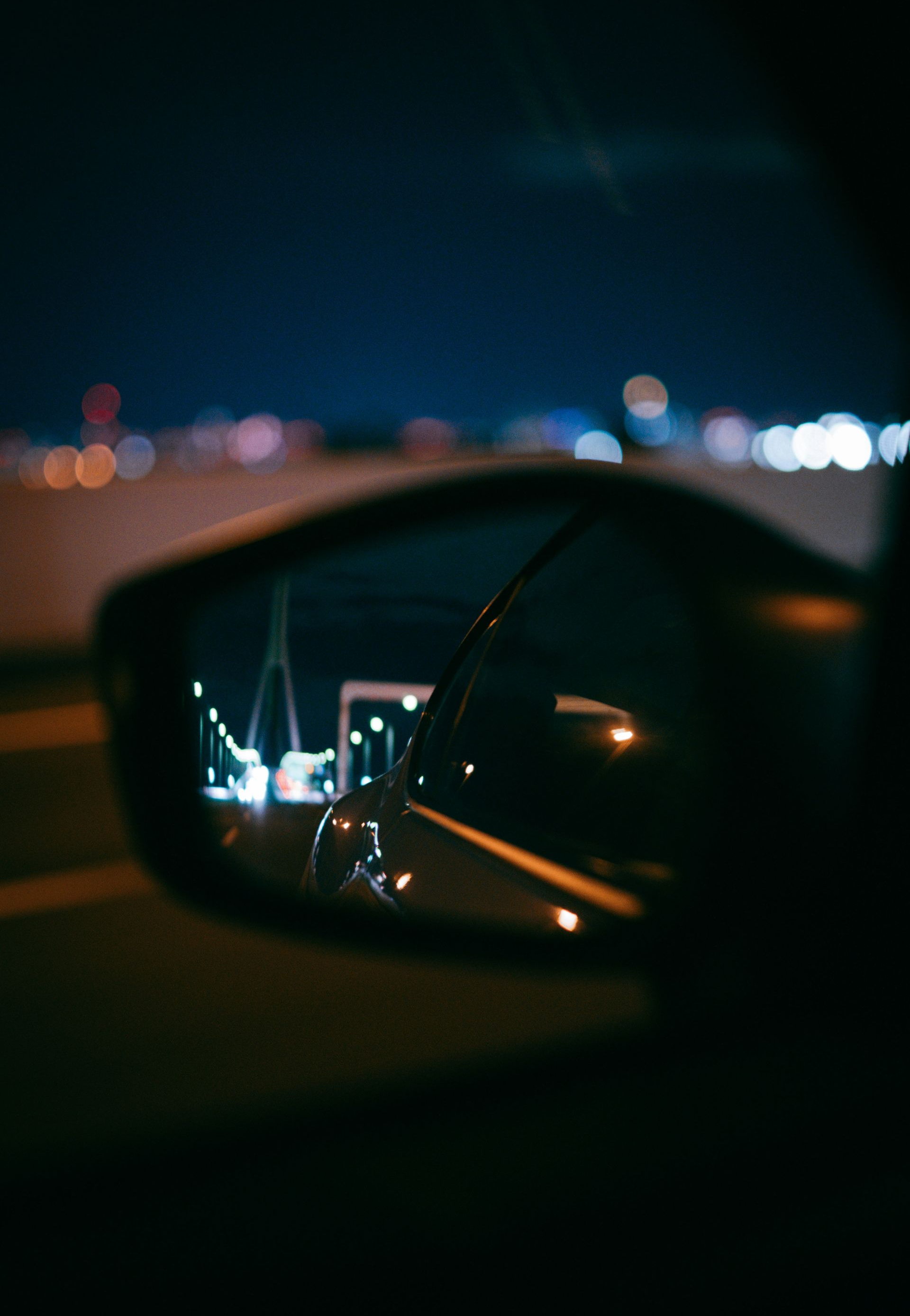 Car's side mirror reflecting a nighttime cityscape with bridge lights, against a dark blue sky.
