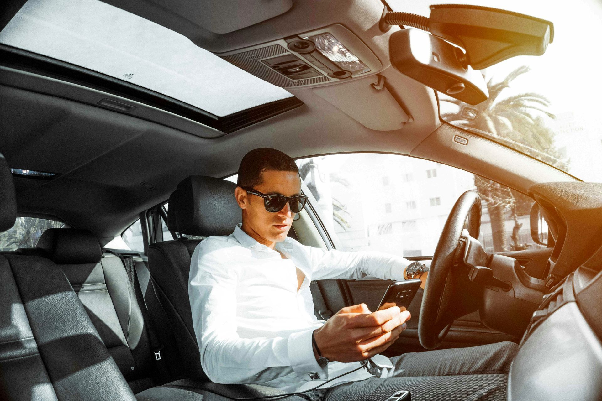 Man in sunglasses, wearing a white shirt, looking at his phone while sitting in the driver's seat of a car.