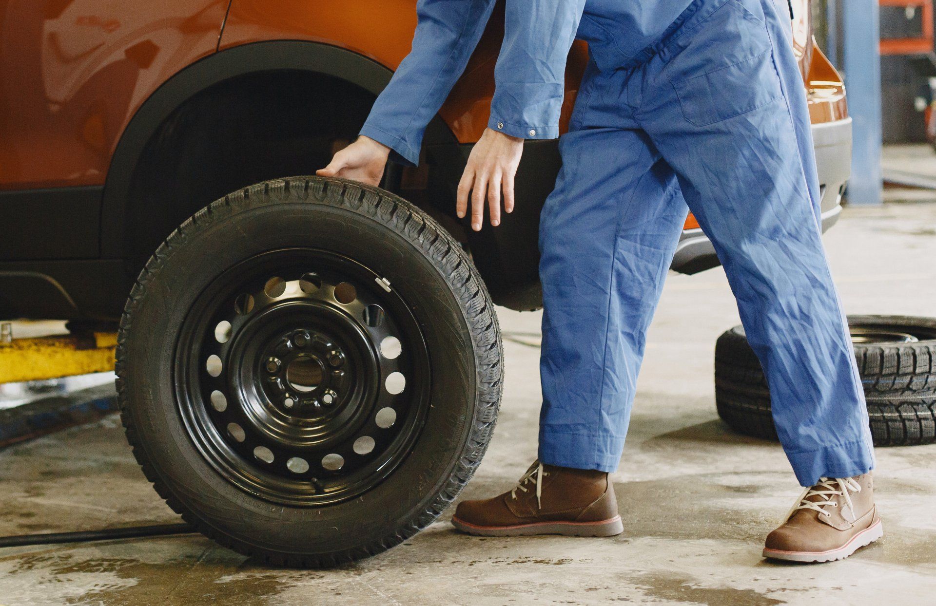 Mechanic in blue overalls replacing a car tire in a garage. He is lifting the new tire near an orange car, with another tire in the background.