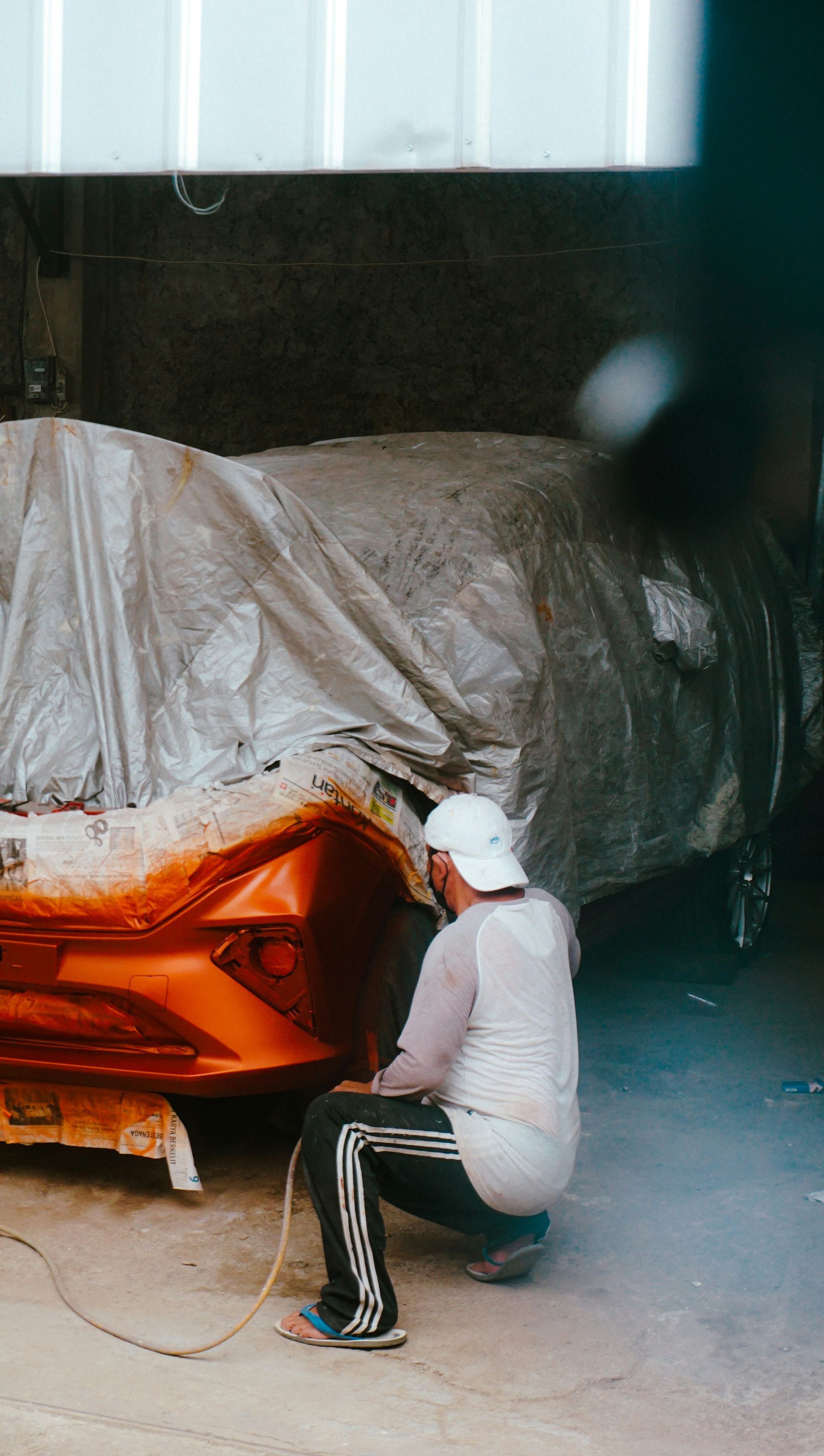 A person in a white shirt and black athletic pants crouches while spray-painting the front bumper of an orange car.