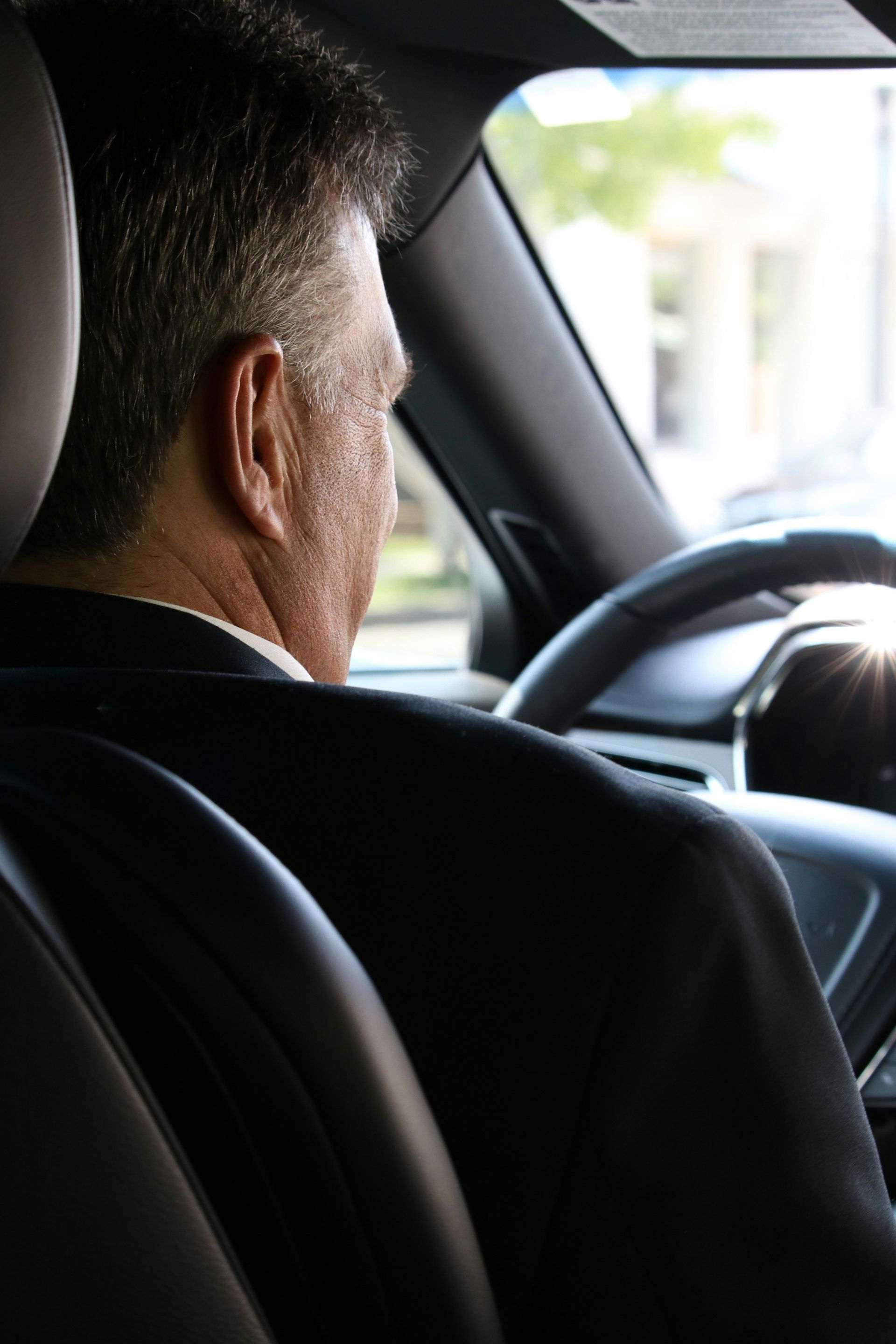 Man in a suit driving a car, seen from the backseat. Sunlit road ahead.
