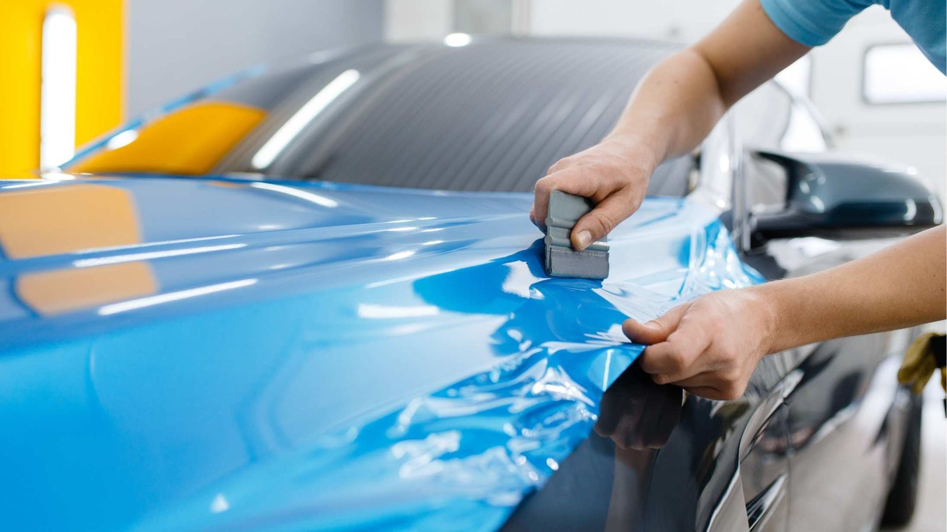 Person applying blue vinyl wrap to the hood of a car with a squeegee in a shop.