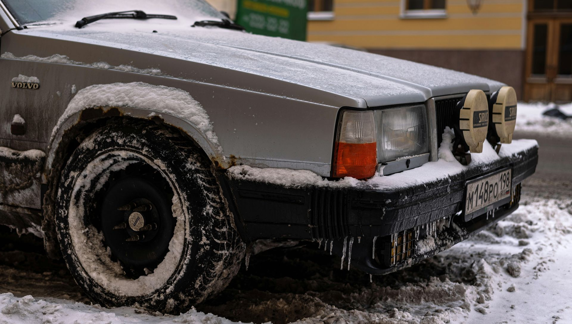 Silver Volvo car covered in snow, parked on a snowy street. Two spotlights on the bumper.