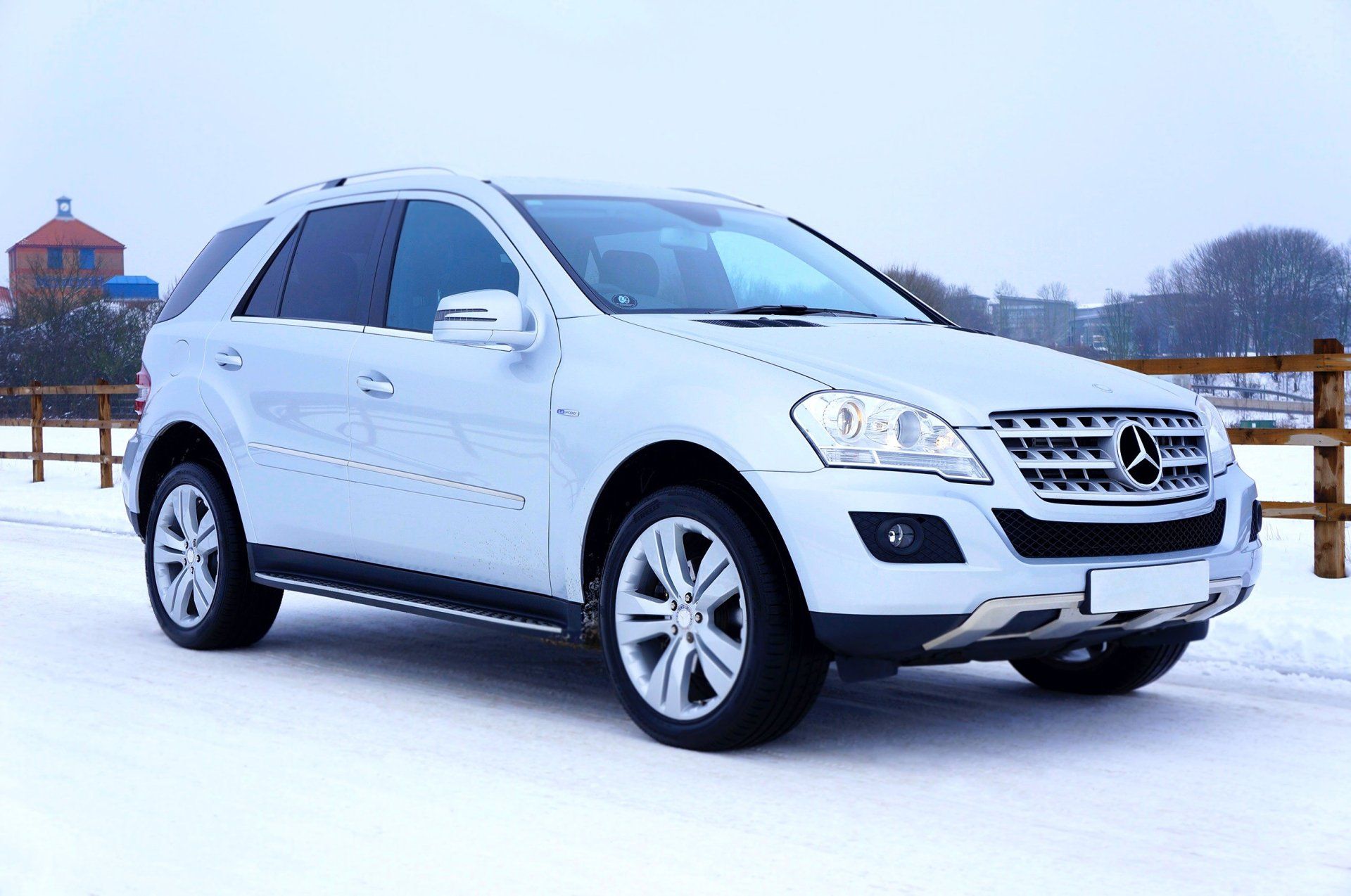 Silver Mercedes-Benz SUV parked on a snow-covered road in front of a wooden fence and a building.