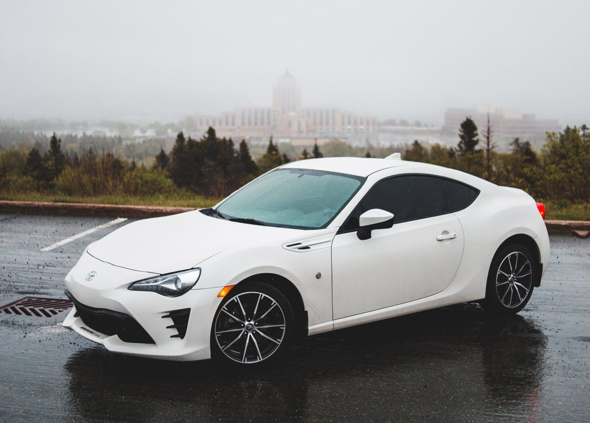White sports car parked on a wet pavement in front of a city on a cloudy day.