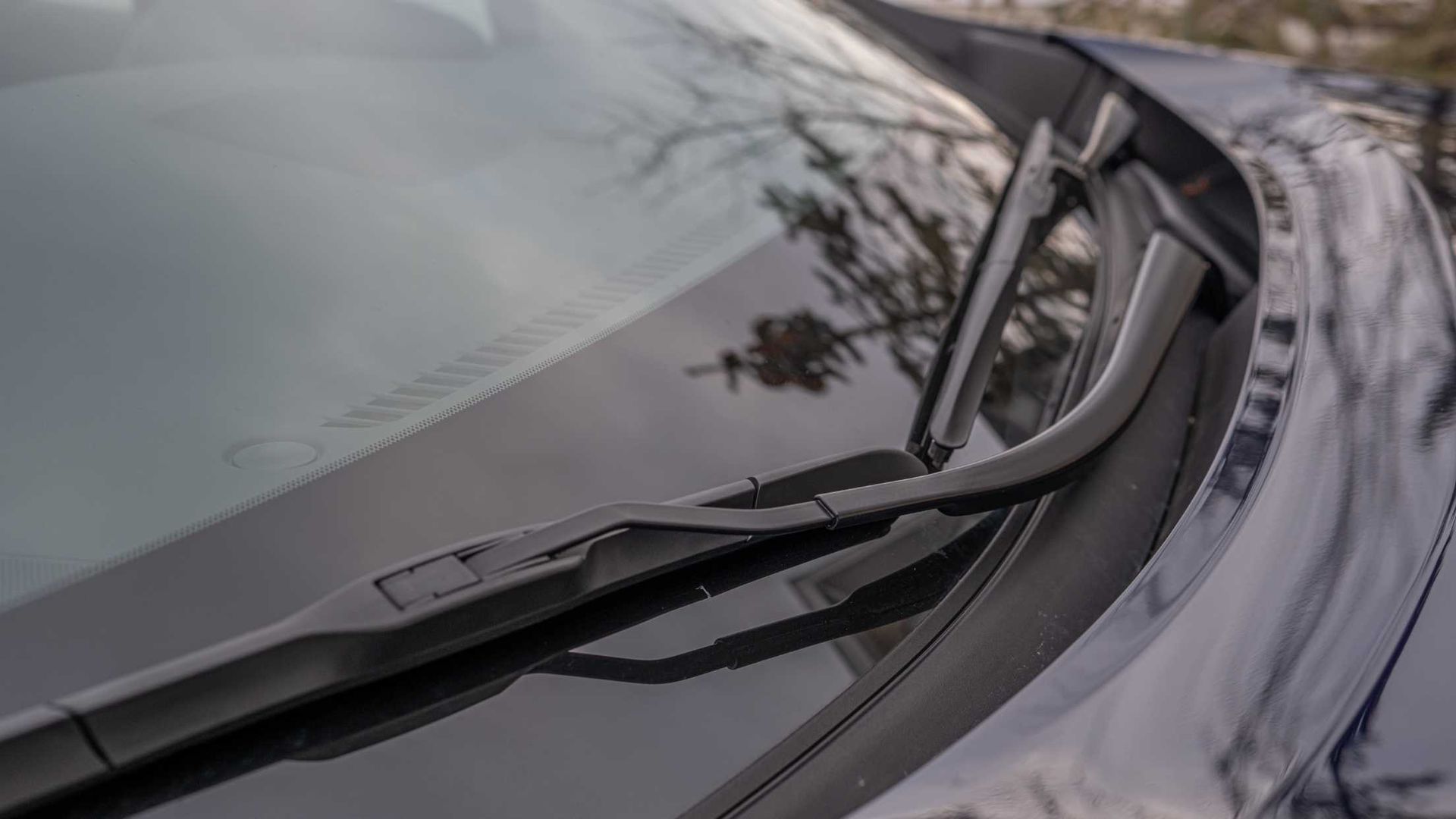 Close-up of a car's windshield with wiper blades in their resting position. The dark blue hood reflects the environment.
