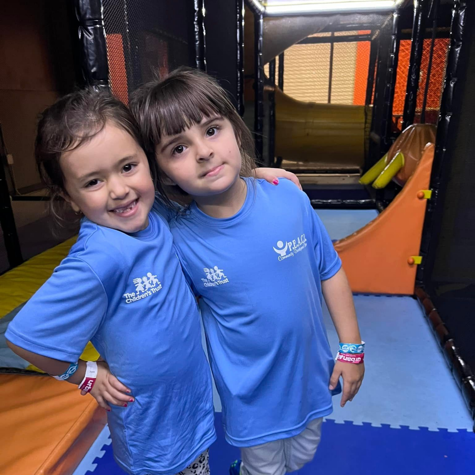 Two young girls in blue shirts hug in an indoor play area with colorful structures.