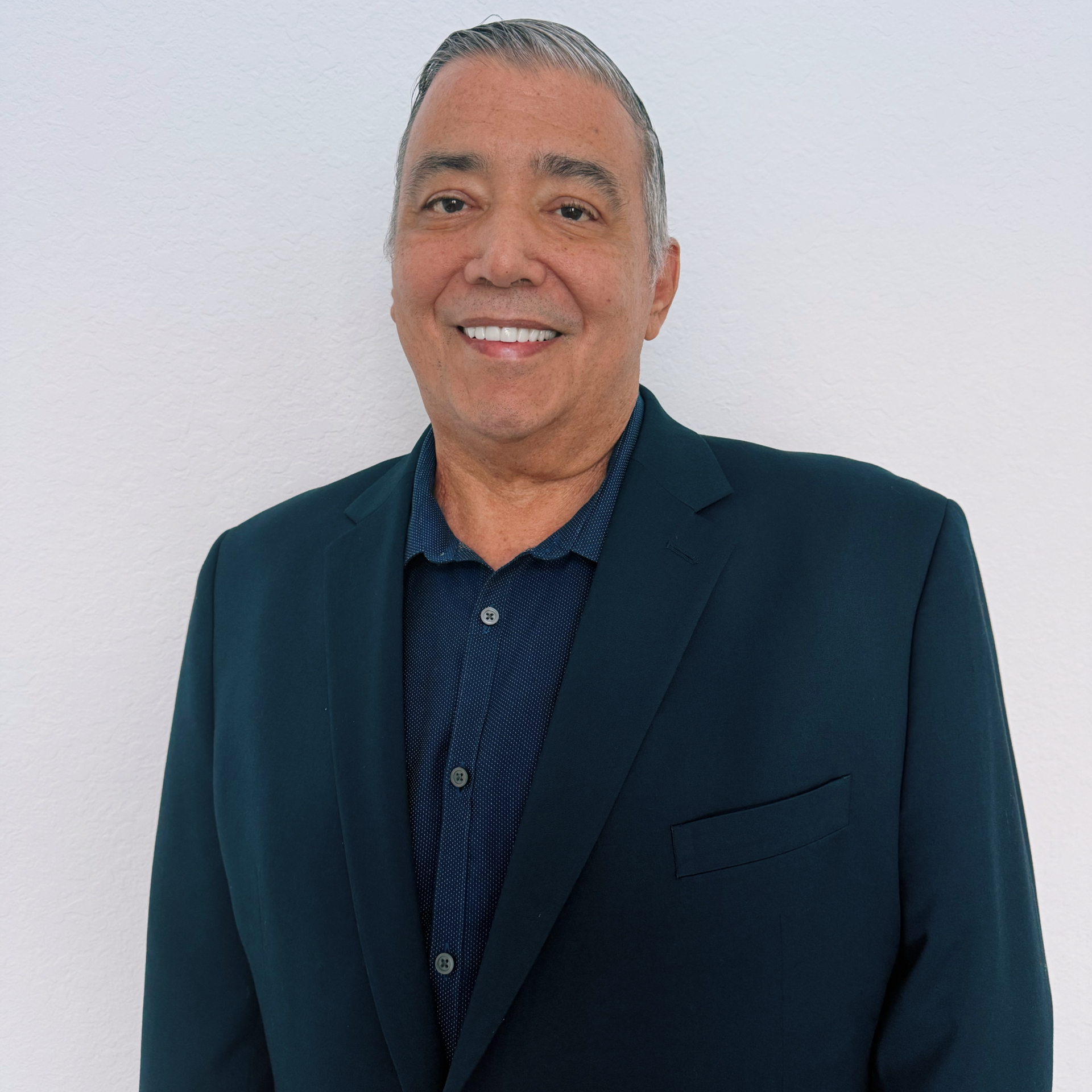 Man smiling in a navy blazer over a blue button-up shirt, against a white wall.