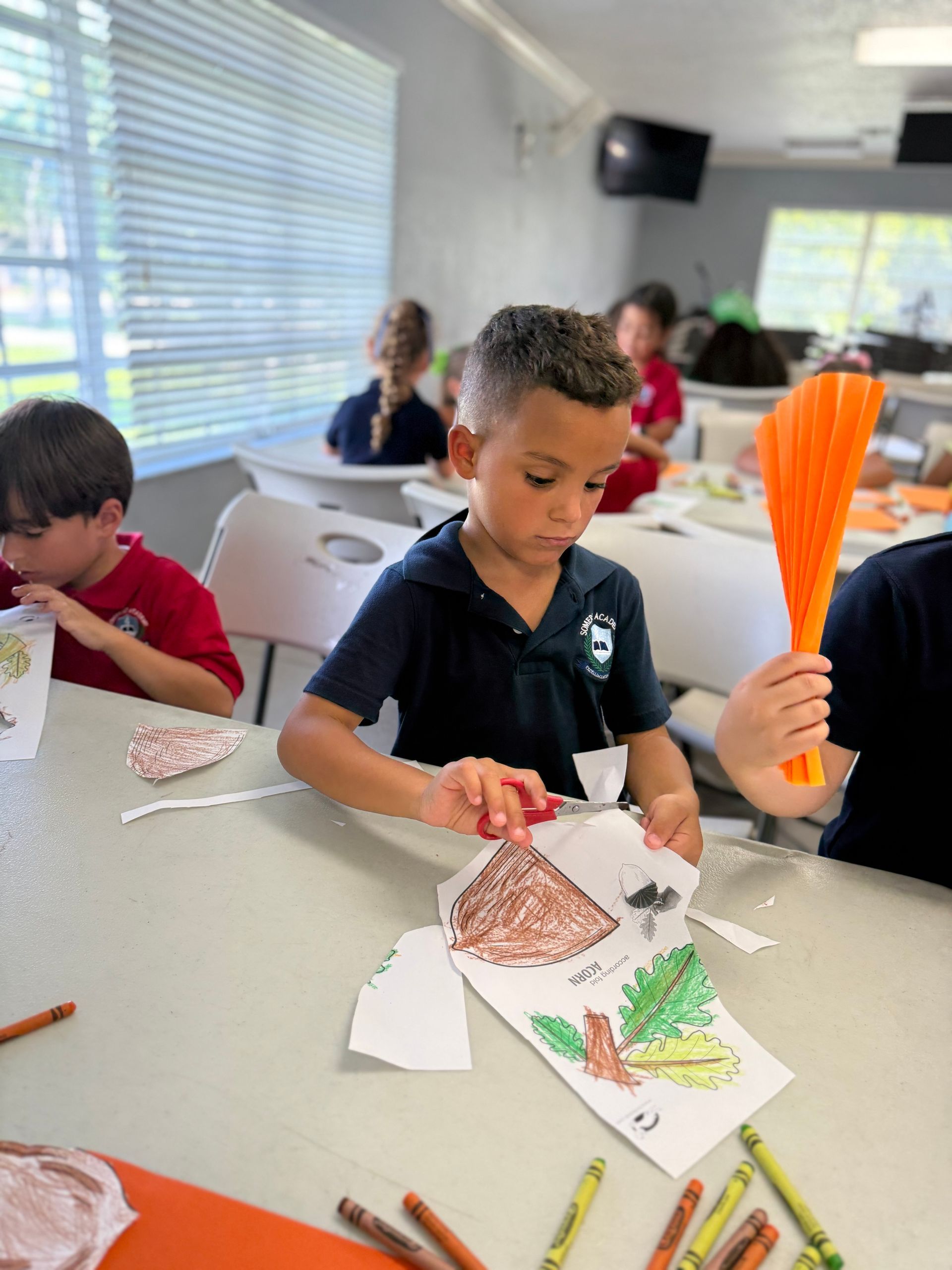 Boy cutting paper at a table, with other children in the background. Colorful crayons are scattered on the table.