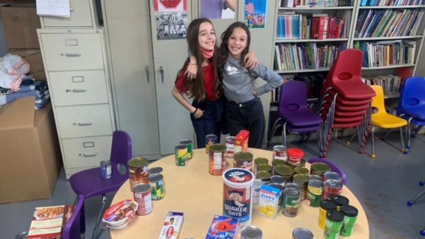 Two people pose with canned goods on a table. Shelves of books, file cabinets, and chairs are in the room.