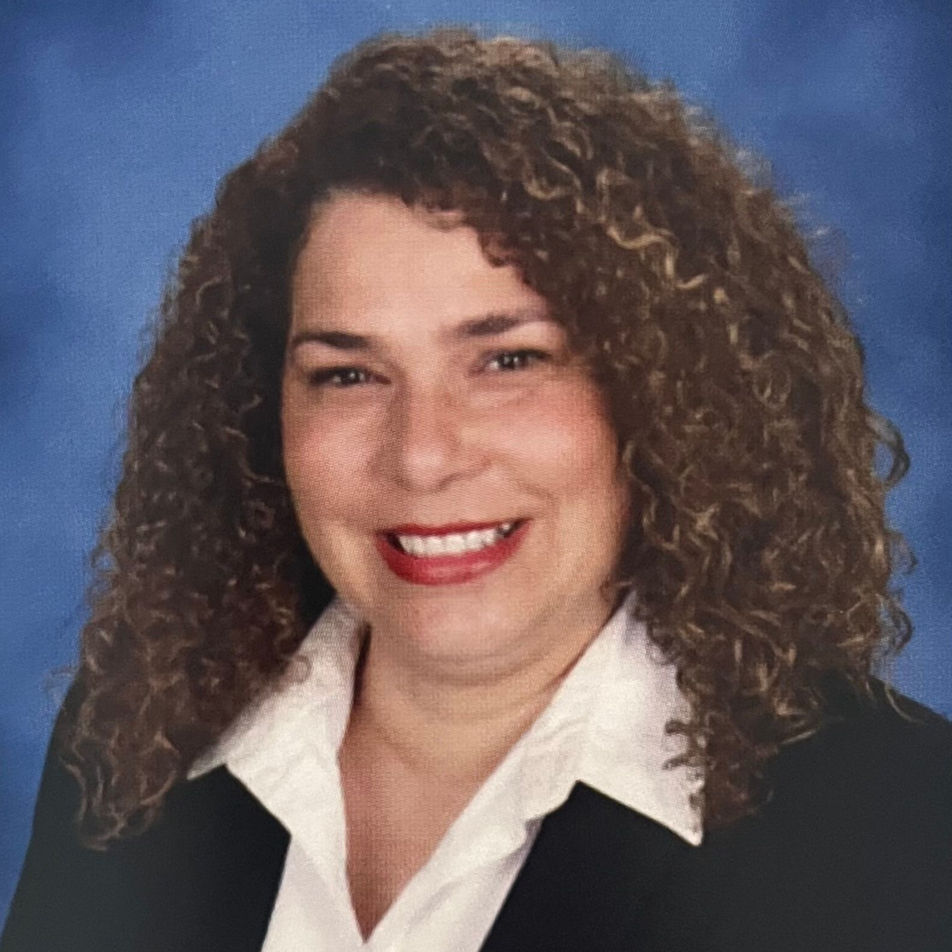 Woman with curly brown hair, smiling, wearing a white collared shirt and black blazer.