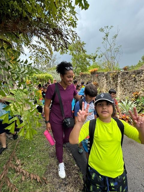 Group walking on path, led by woman in purple scrubs. Boy in neon shirt makes peace sign. Outdoors, cloudy sky.
