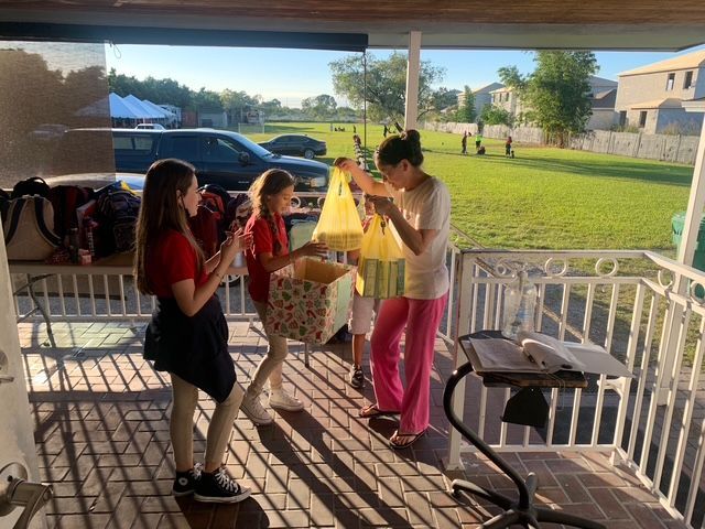 Three young people on a porch, handing out bags near a basket. Sunlight streams across the scene.