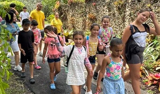 Group of people walking on a path in a garden. Some children wave and smile, surrounded by plants.