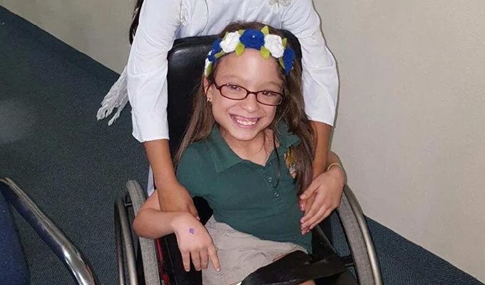 Girl in a wheelchair, smiling with a flower crown, is being pushed by someone in a white coat.