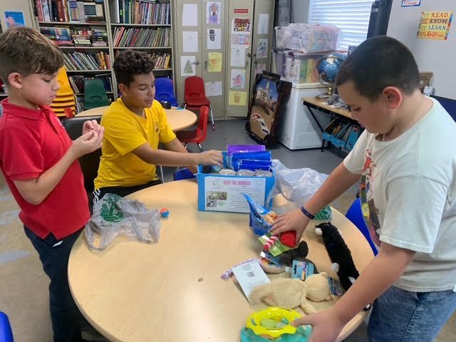 Three children sorting items at a round table in a classroom with bookshelves.