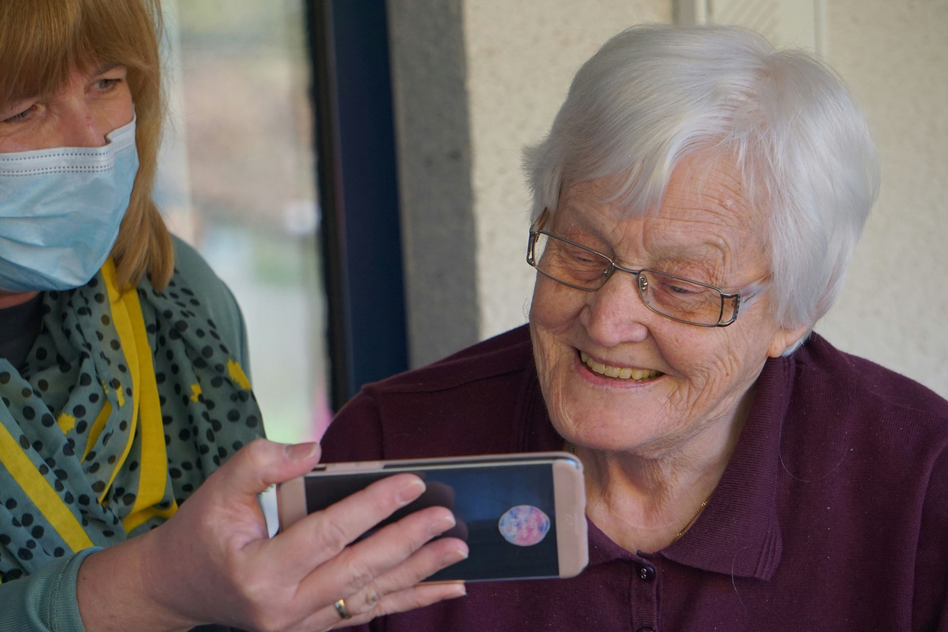 A woman wearing a mask is helping an elderly woman use a cell phone.